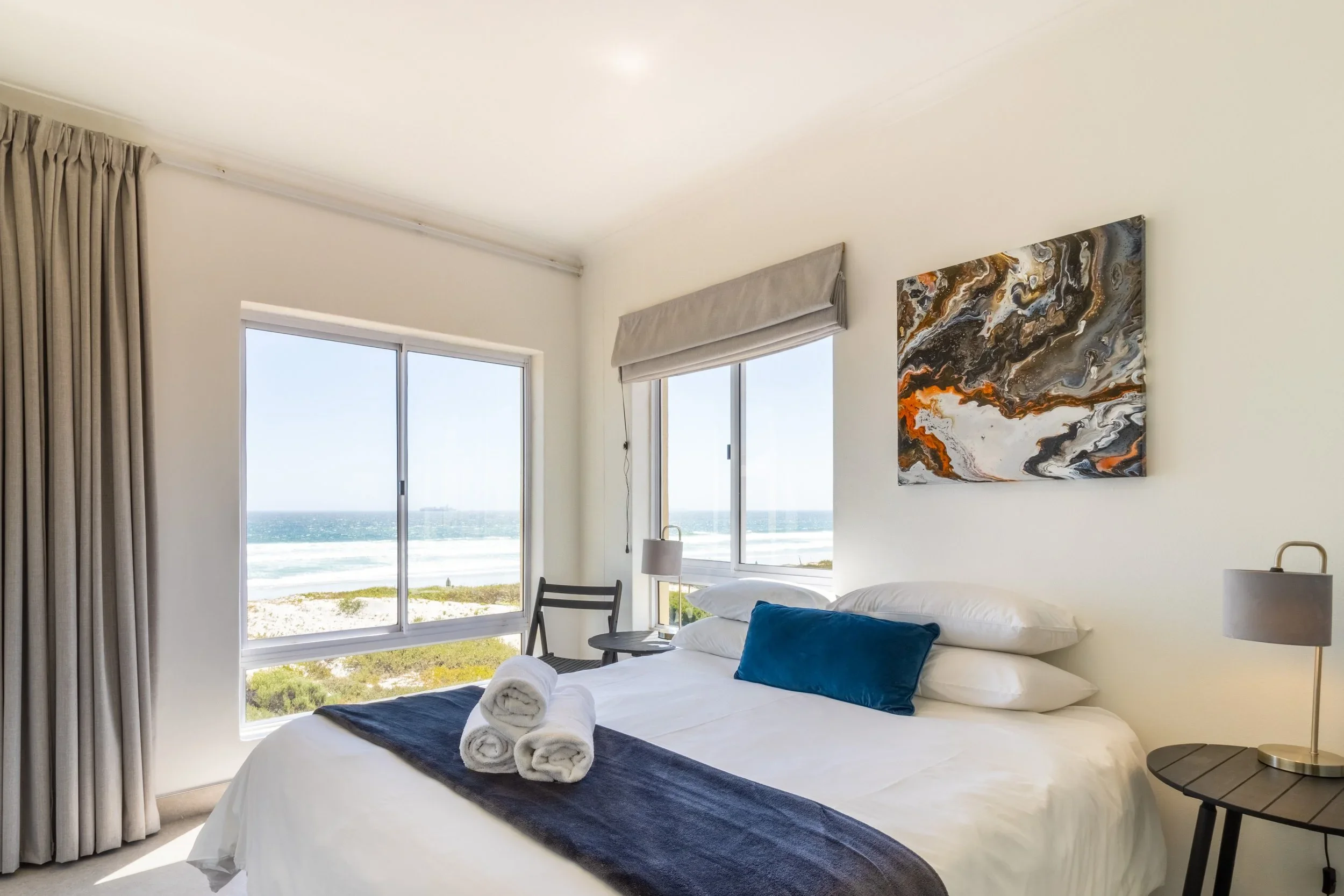 A bedroom with white bedding, a navy blue blanket, and three rolled towels on the bed; large windows with ocean view, abstract wall art, beige curtains, and a small black chair beside a window.