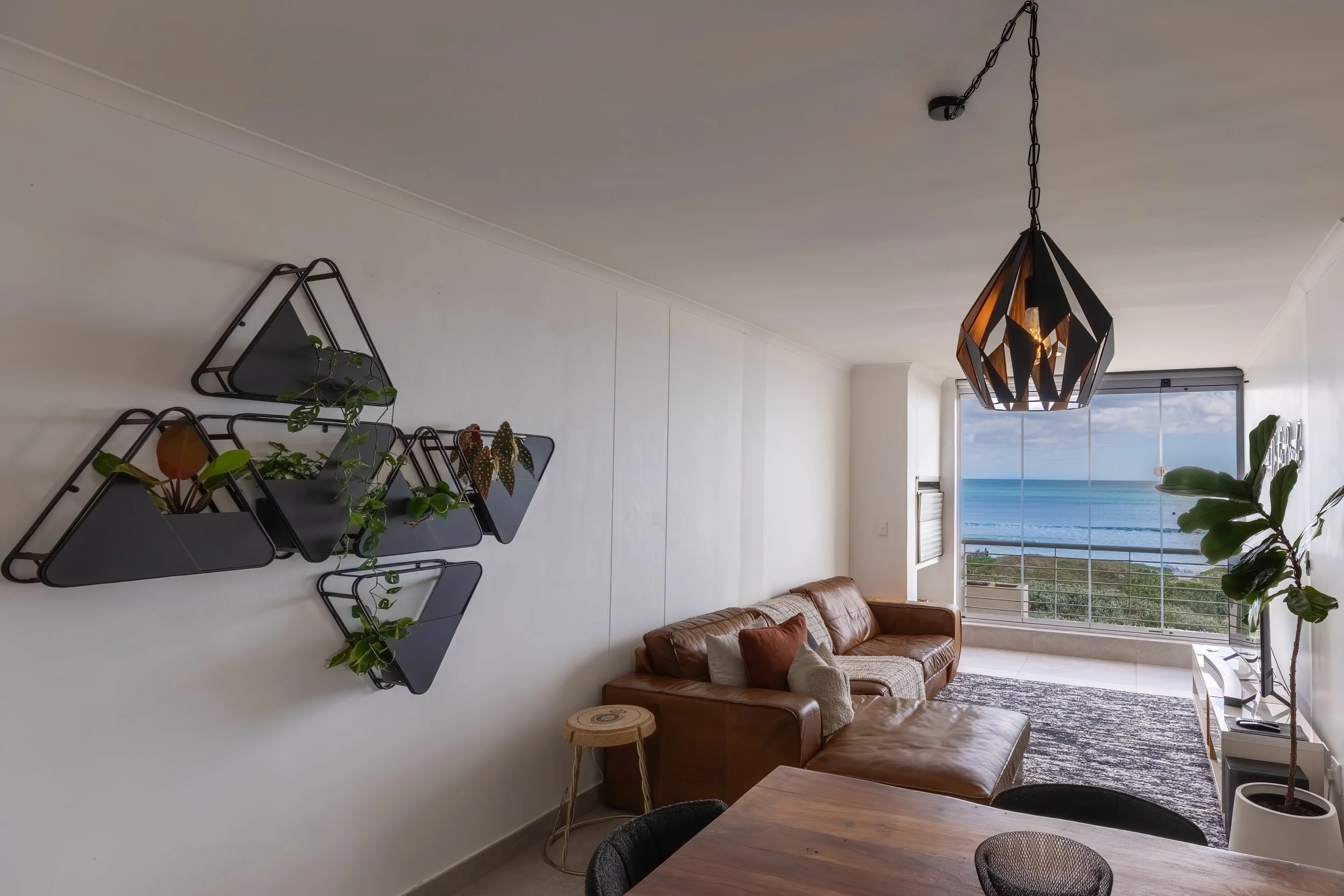 Living room with a brown leather sofa, a wooden dining table, black chairs, indoor plants, modern black geometric pendant light, and a large window with a view of the ocean and sky.