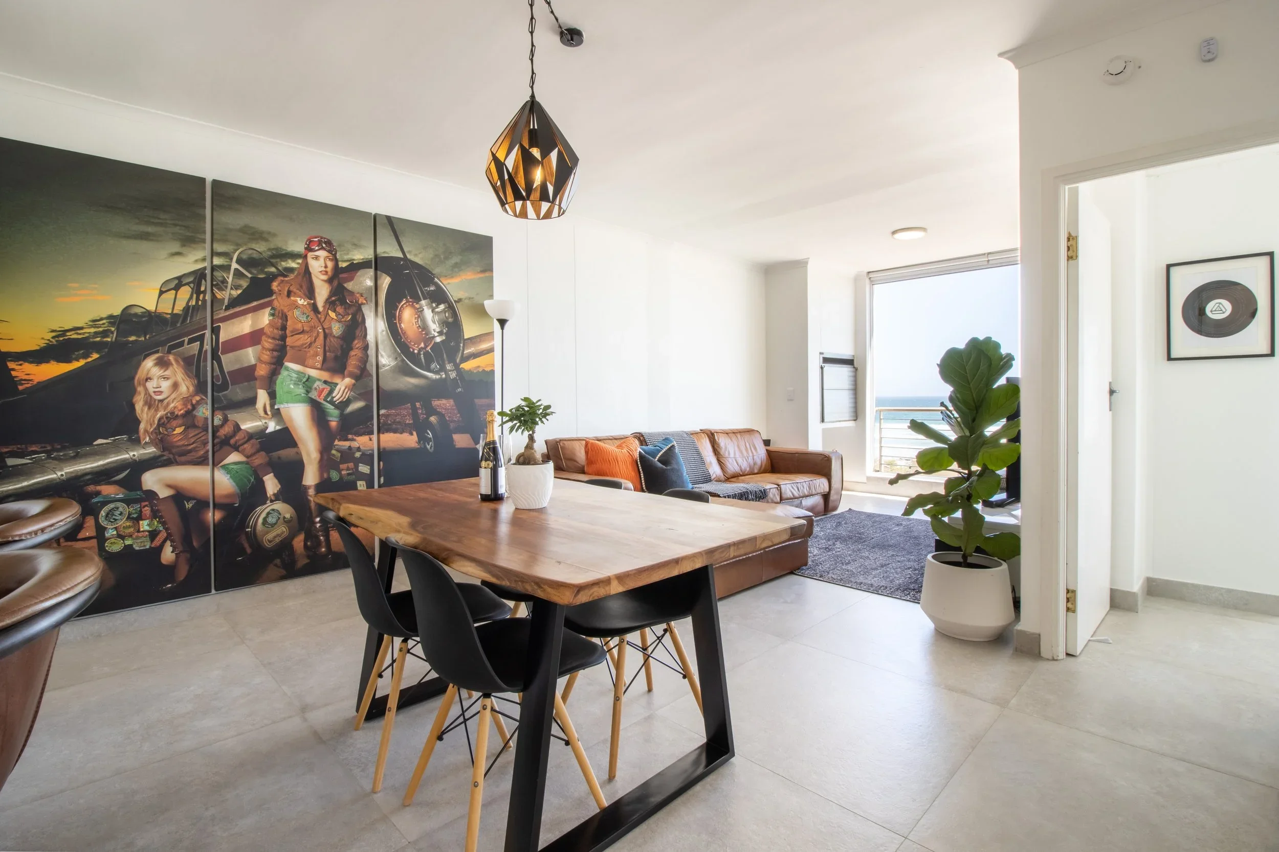 Living room with a large wood dining table, black chairs, a brown leather couch, green plant in a white pot, large windows with a view of the ocean, and a black and white framed picture of a vinyl record hanging on the wall.
