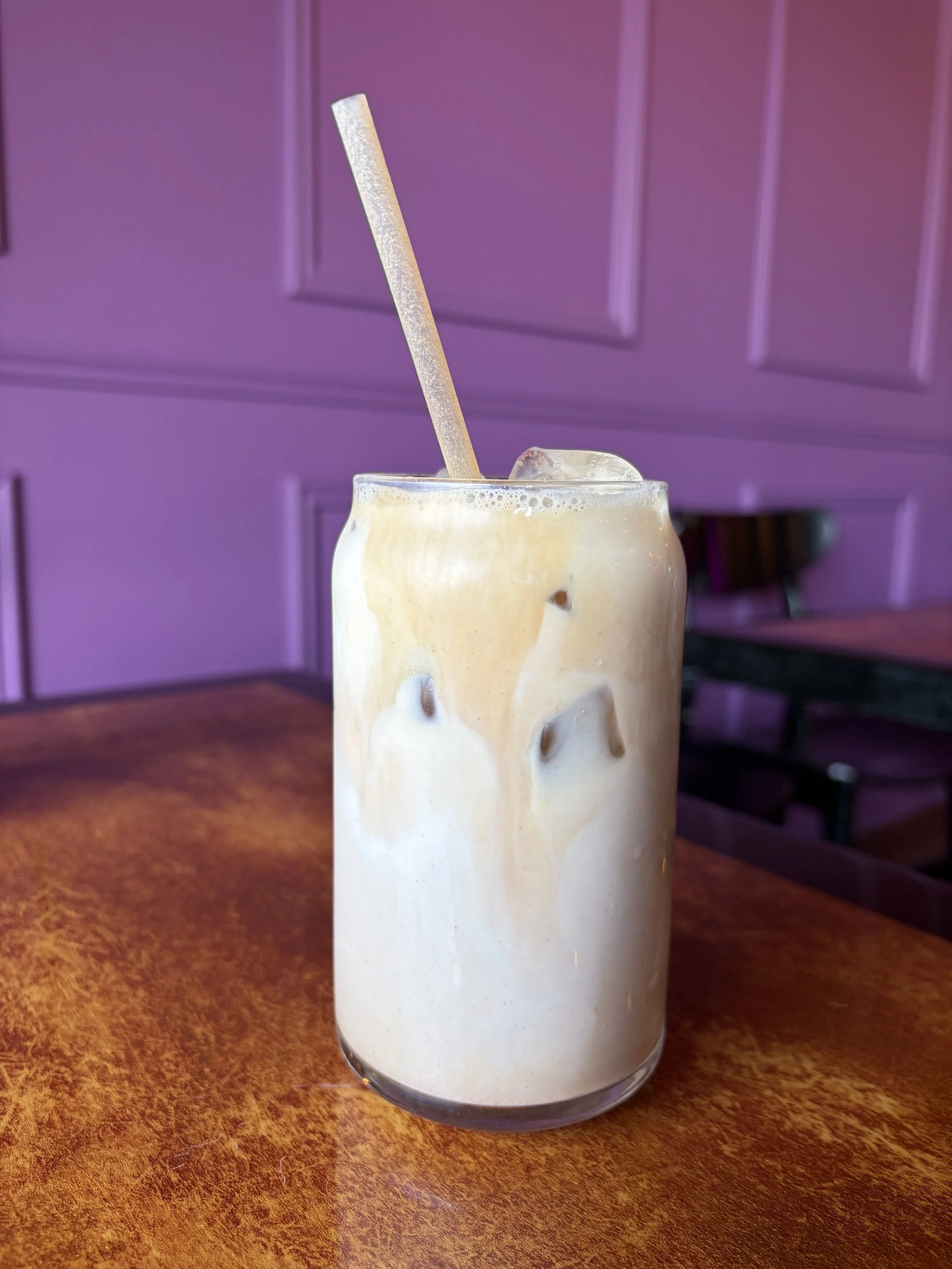 A tall glass of iced coffee or latte with a straw, sitting on a wooden table with a purple paneled wall in the background.