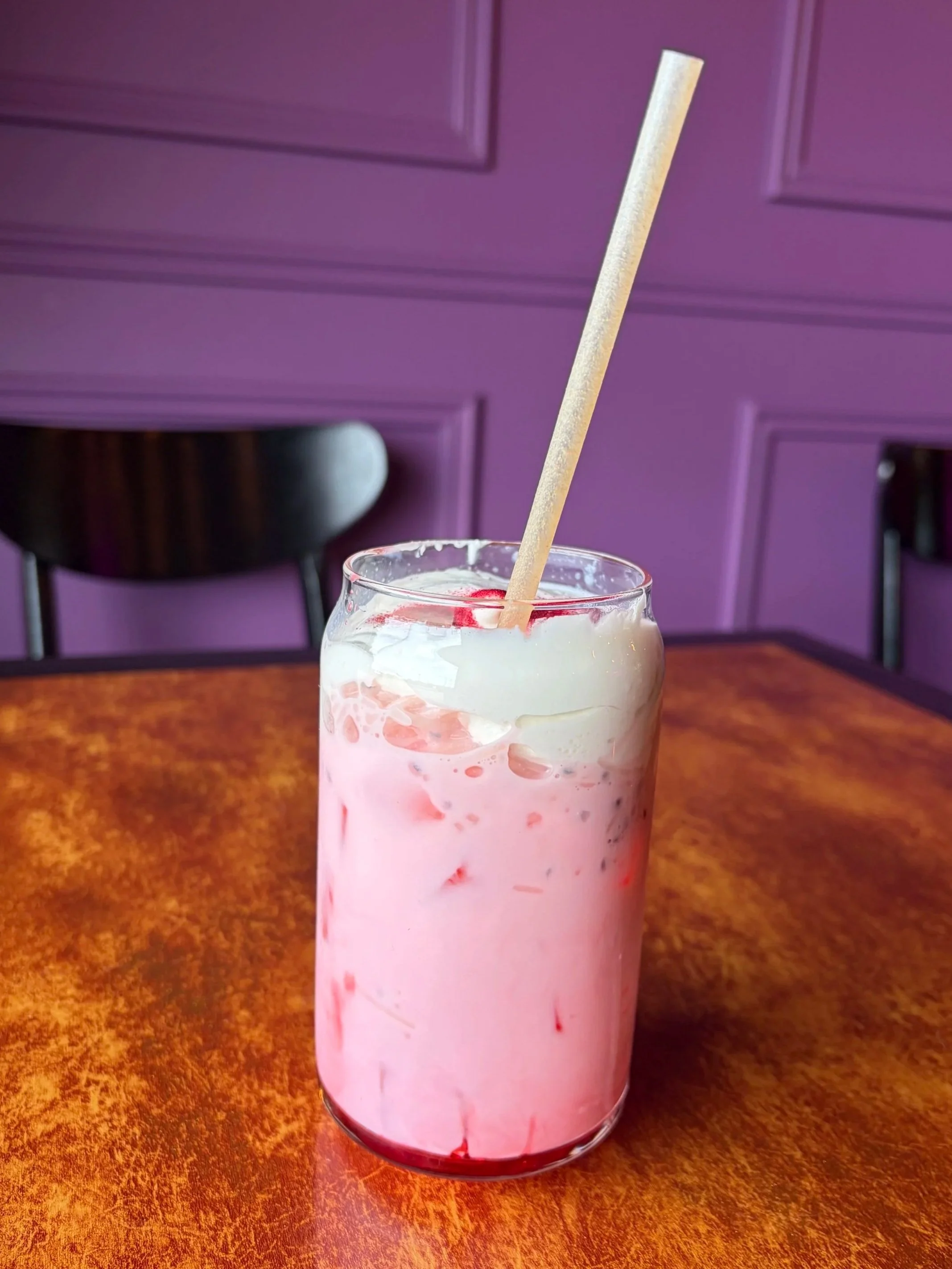A tall glass of pink milkshake with red syrup at the bottom, served on a wooden table, with a purple wall in the background.