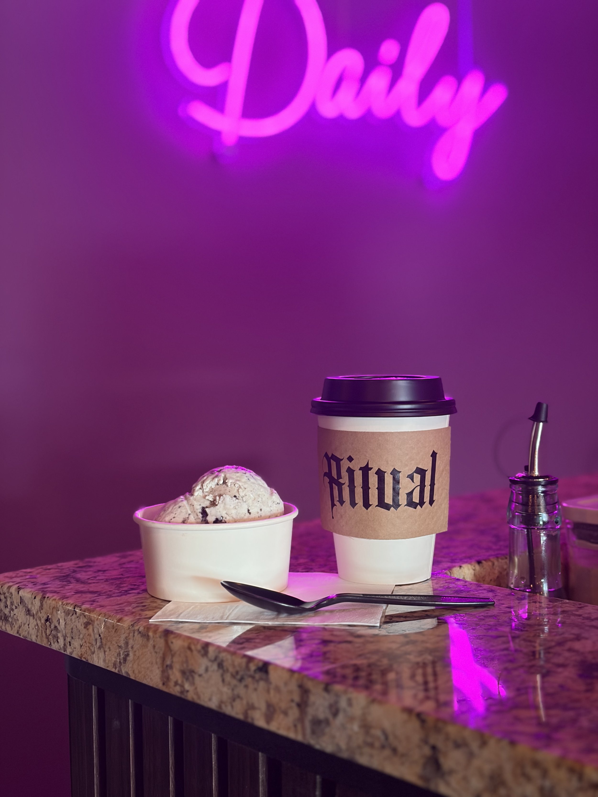 A cup of ice cream with chocolate chips, a to-go coffee cup with a brown sleeve that says 'Digital', and a small glass jar with a metal straw on a granite countertop with a purple neon sign in the background.