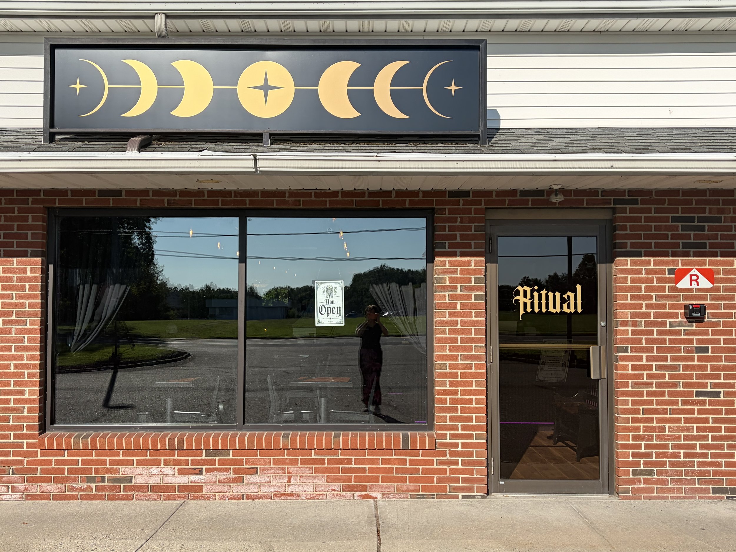 Storefront with a sign featuring phases of the moon and a star, and a window with a sign that reads 'Now Open', brick and siding exterior, glass door, and reflection of trees and power lines.