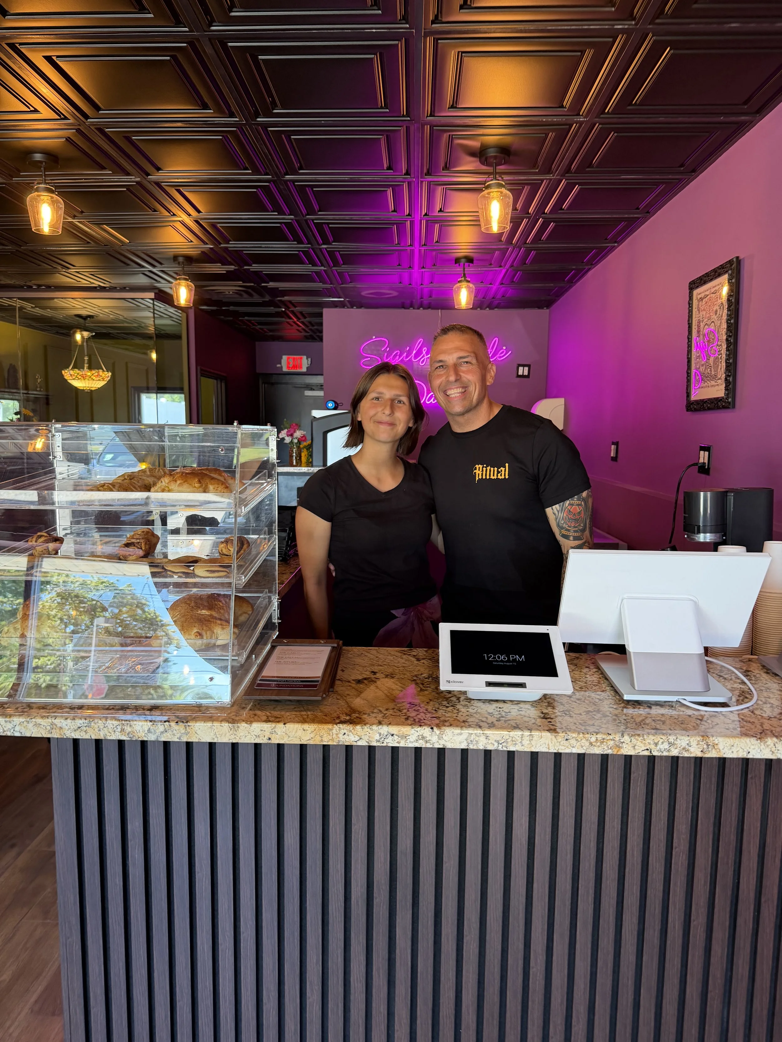 A man and a woman standing behind a bakery counter with a bakery display case filled with croissants, danishes, and pastries. The background features a neon sign that reads 'Sleight' and purple lighting.