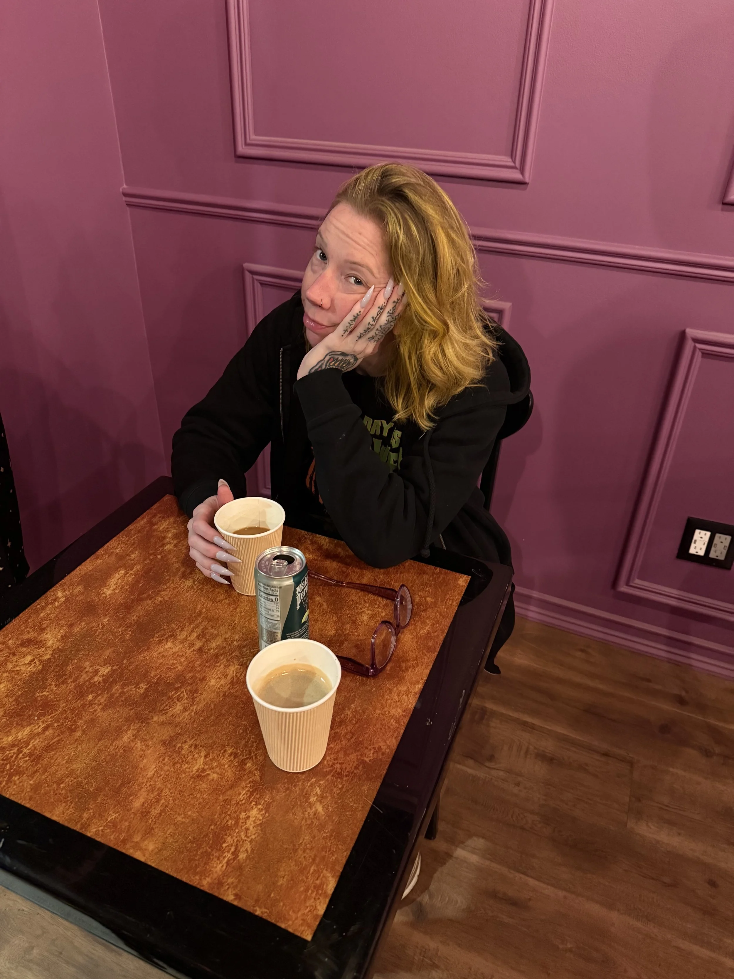 A woman with blonde hair and tattoos on her hand is sitting at a wooden table with two coffee cups, a can, and glasses, against a pink wall with decorative molding.