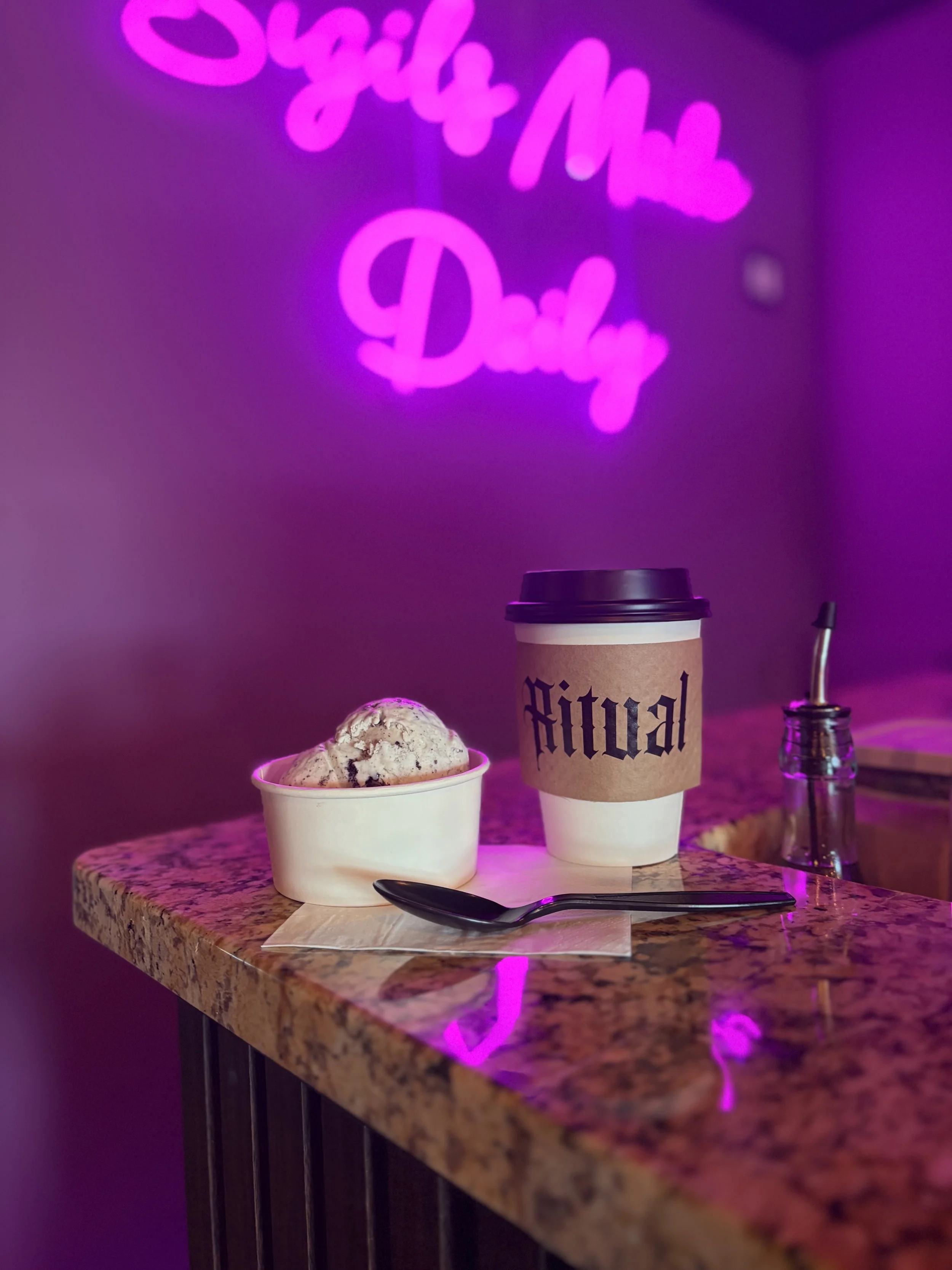 A cup of ice cream on a marble table with a paper coffee cup labeled 'Ritual' behind it, a small glass container with a metal straw, and a black spoon on a napkin. In the background, a neon pink sign reads 'Sweet & More Daily' and illuminates the scene with a purple hue.