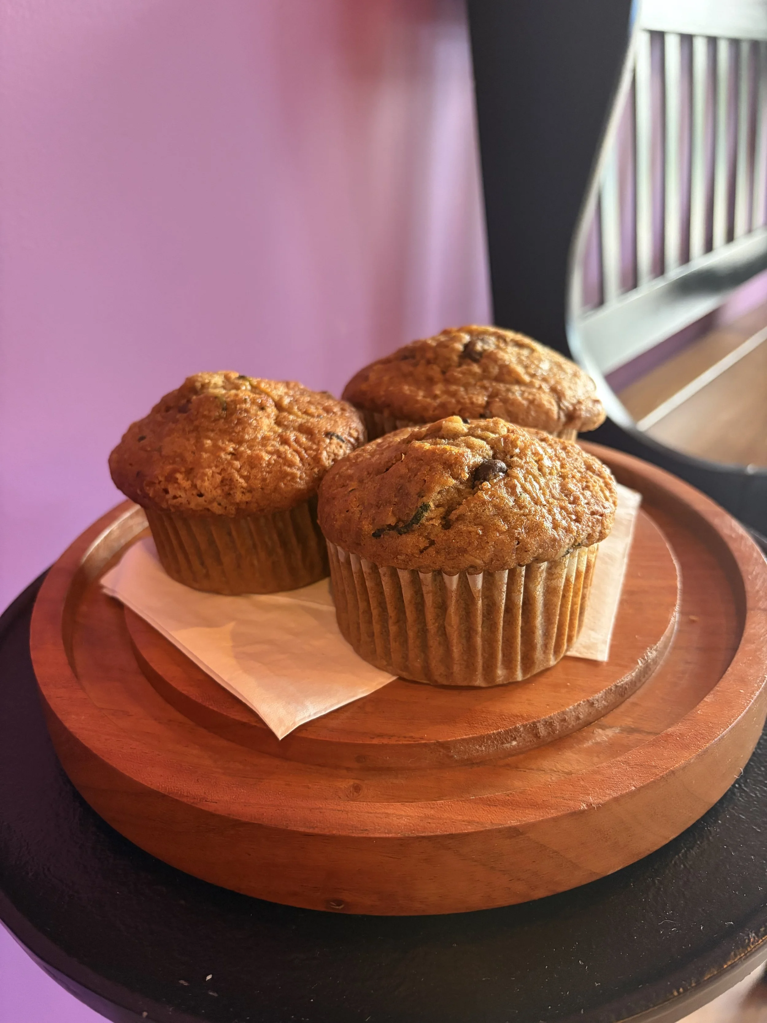 Three muffins on a wooden plate with a paper liner, placed on a black table with a purple wall in the background.