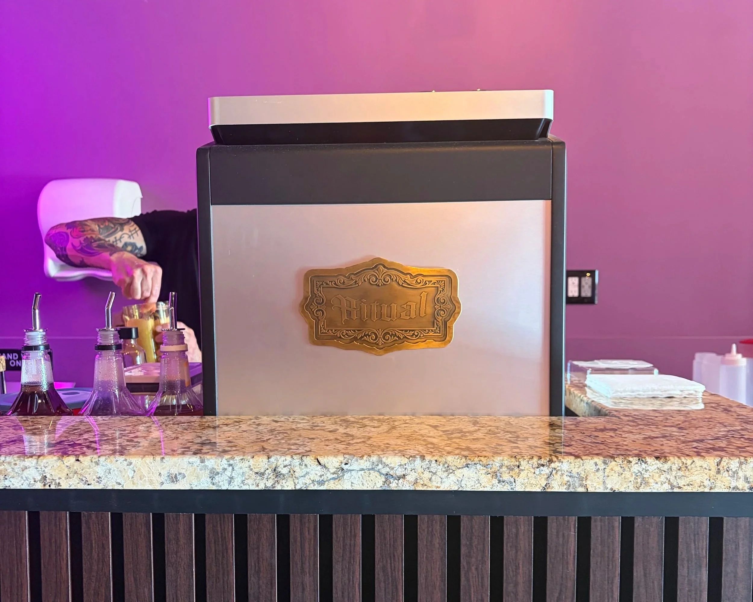 A bar counter with glasses and bottles, a large metallic beverage cooler with a gold plate labeled "Royal", and partial view of a person with a tattooed arm wearing a black shirt. The background wall is painted purple with colorful lighting.
