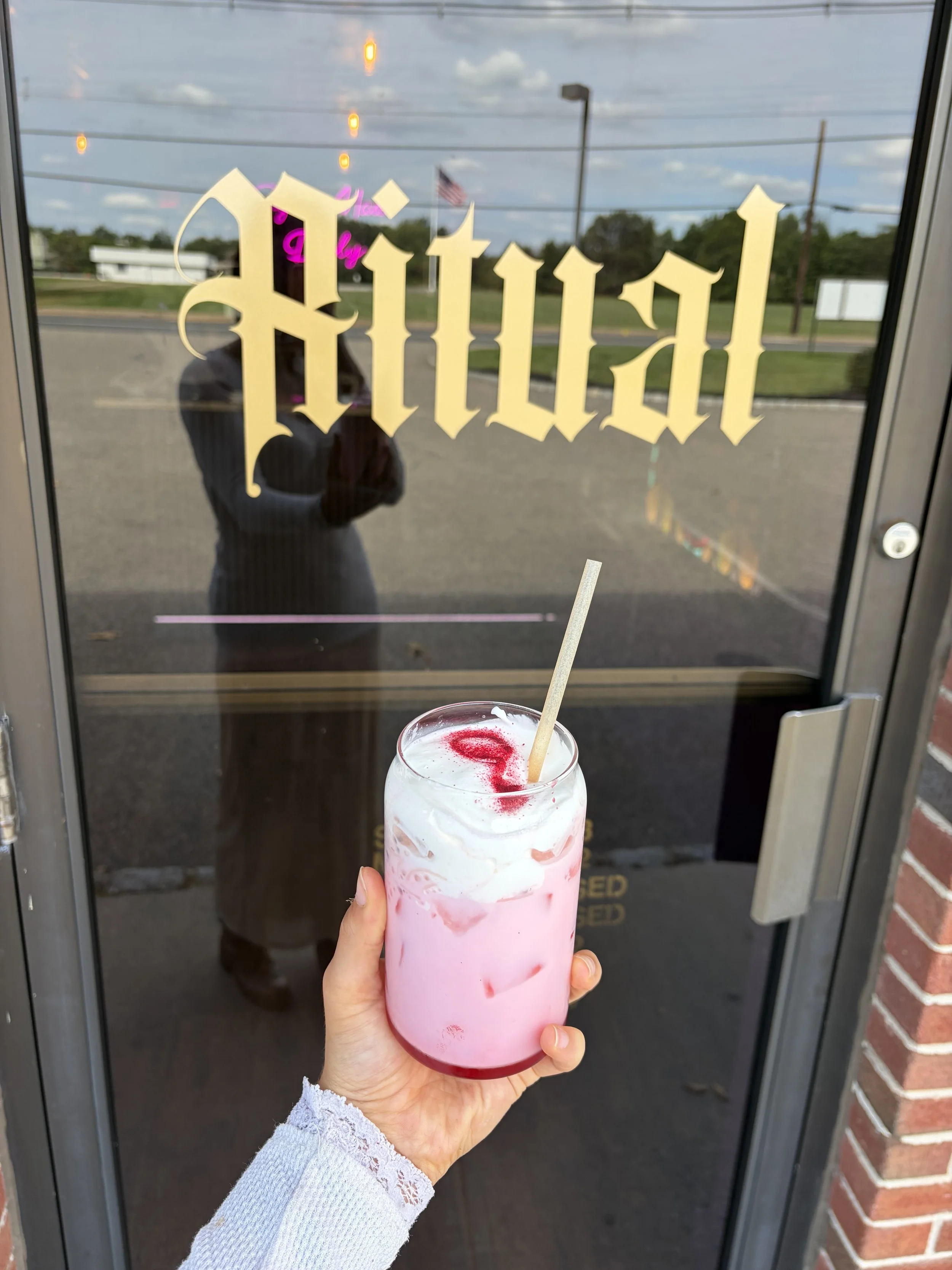 A person holding a pink and white milkshake with a cherry syrup swirl and a straw, standing in front of a glass door with the word 'Open' written in gold Gothic font. The door reflects the person's silhouette, a parking lot, and an American flag in the background.
