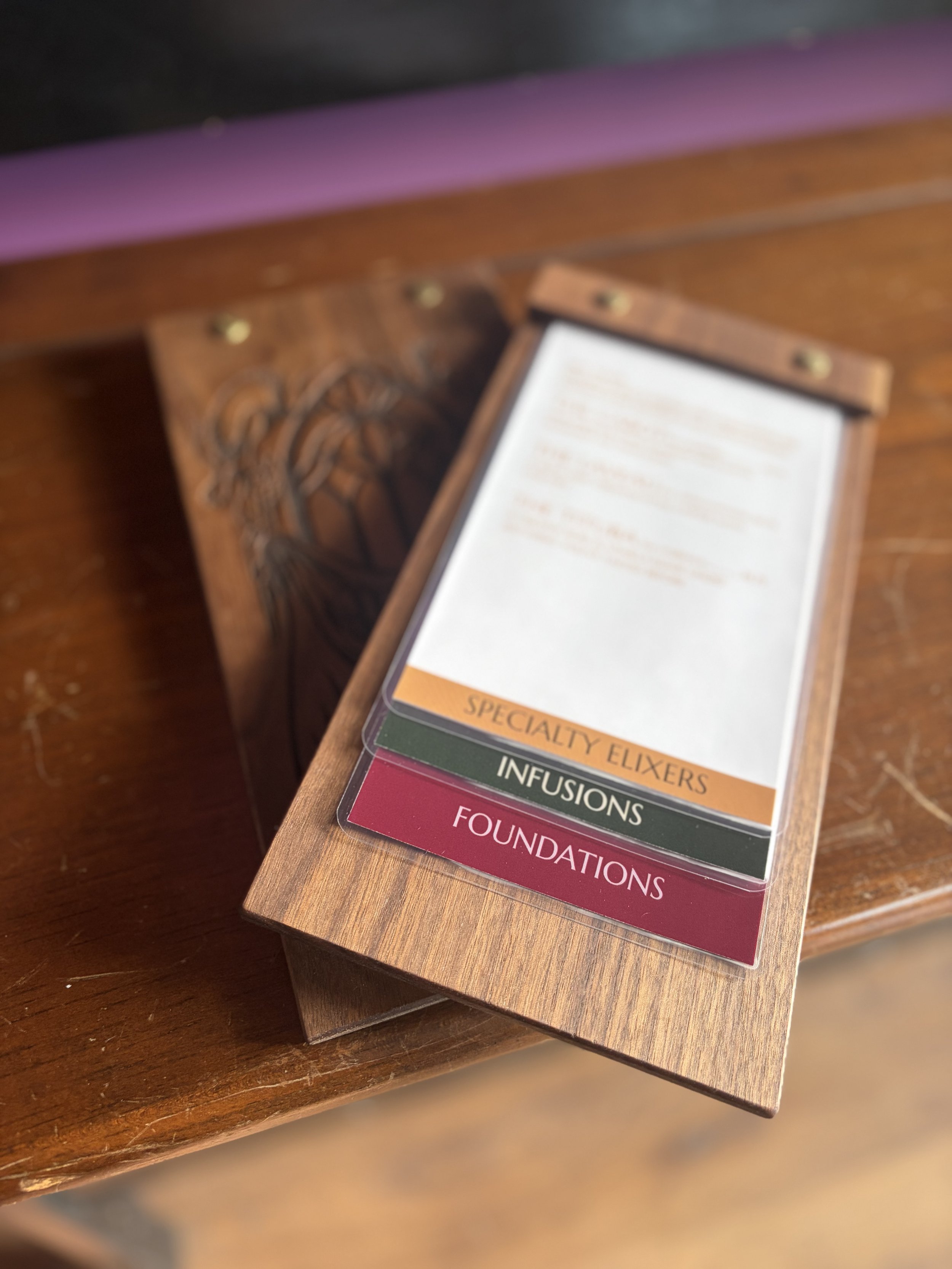 Close-up of a wooden menu holder with a list of wine categories including Foundations, Infusions, Specialty Elixirs, and more, placed on a wooden table.