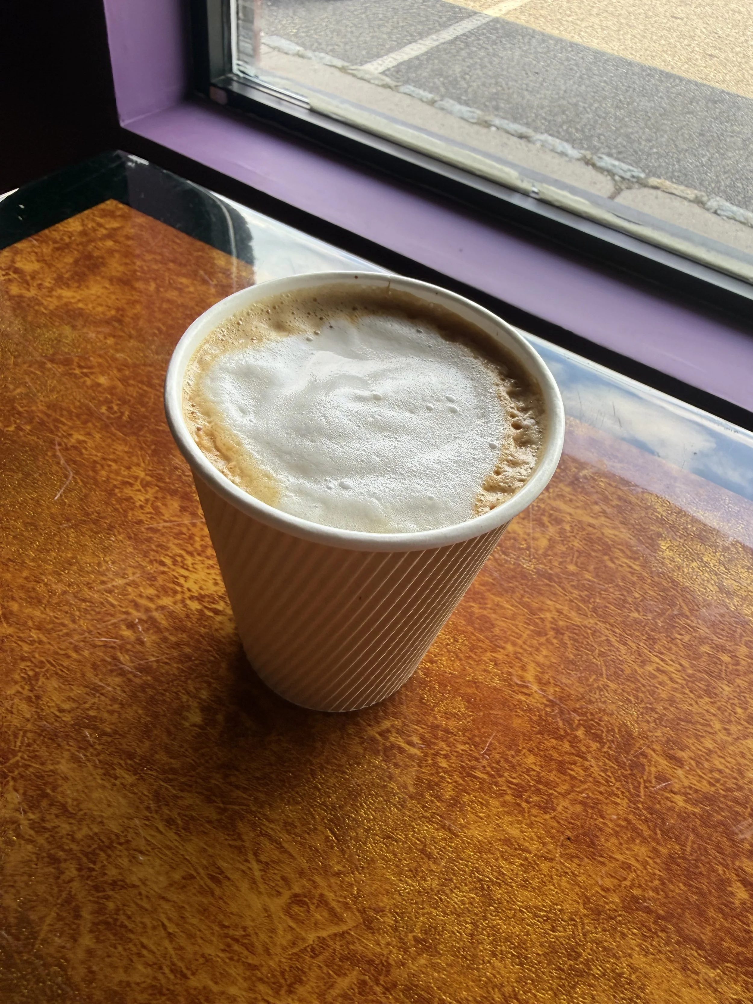A paper cup of coffee with foam on top placed on a wooden table next to a window, with the street visible outside.