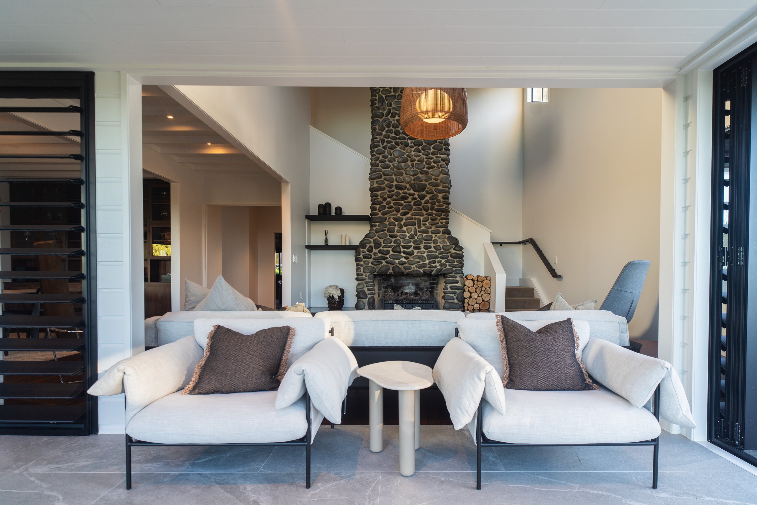 Living room with white sofa, dark brown and black throw pillows, small white coffee table, stone fireplace with logs, ceiling pendant light, staircase, and windows with blinds.