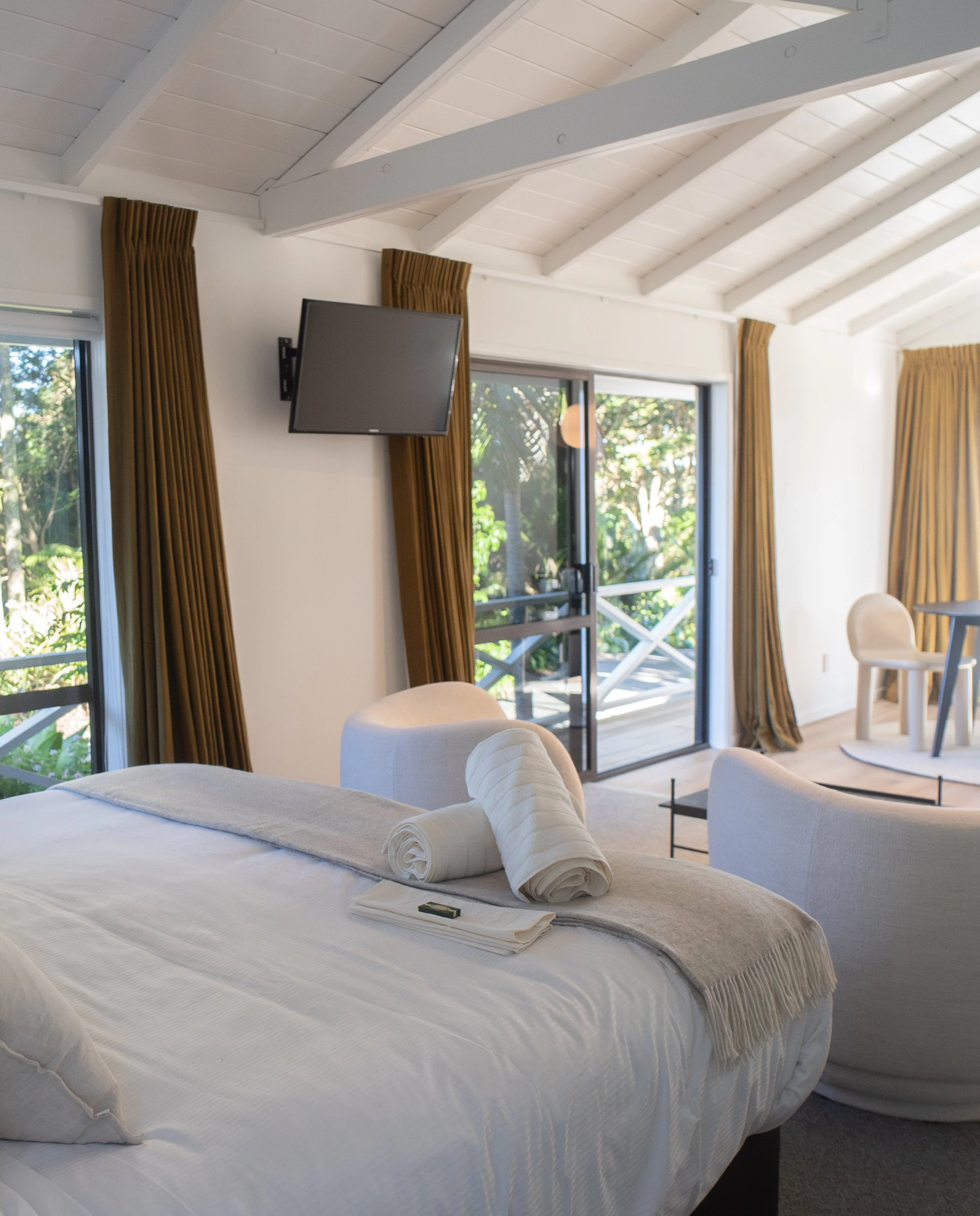 A bedroom with a large bed covered with a white bedspread, accompanied by two rolled white towels and a blanket, placed near a window with brown curtains. The room has a sloped white ceiling with exposed beams, and a small flat-screen TV mounted high