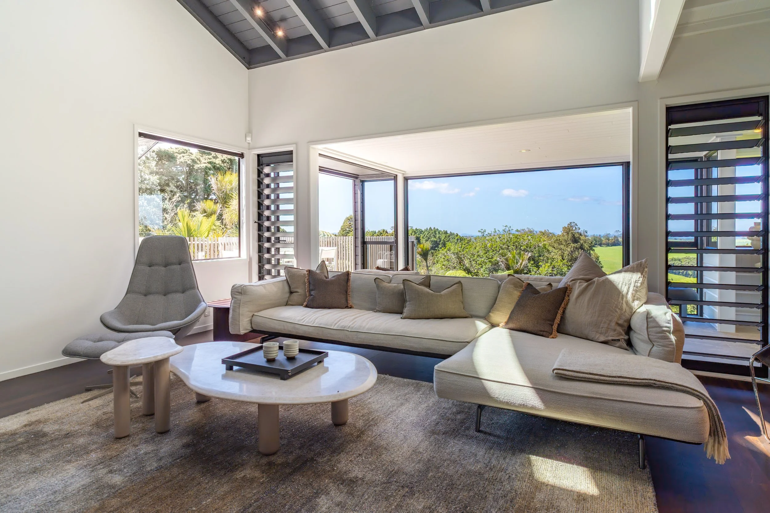 Bright living room with large windows revealing a view of trees and a fence outside. There is a curved beige sofa with multiple pillows, a modern gray chair, round and rectangular coffee tables, and a dark natural fiber rug. The ceiling is vaulted wi