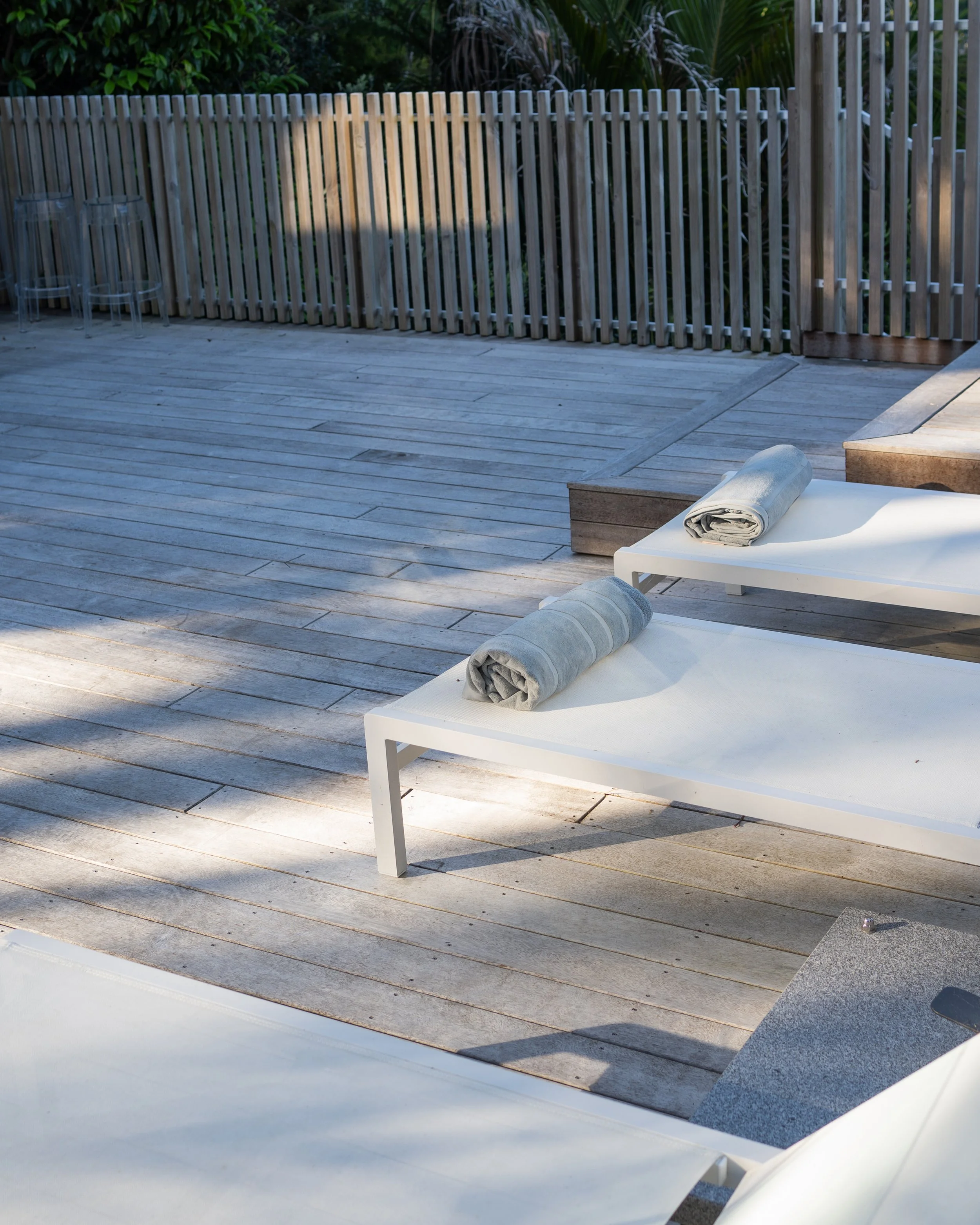 Outdoor deck area with a wooden privacy fence, patio furniture, and a lounge chair with rolled towels, surrounded by greenery.