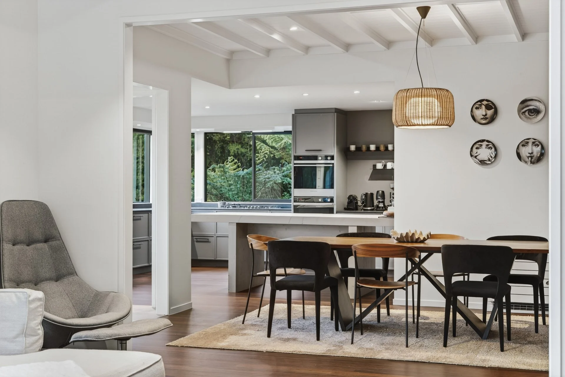 A modern open-concept kitchen and dining area with white walls, a wooden dining table, six black chairs, and a pendant light. There are wall art circular mirrors with black-and-white photos. The kitchen has gray cabinets, built-in appliances, and lar