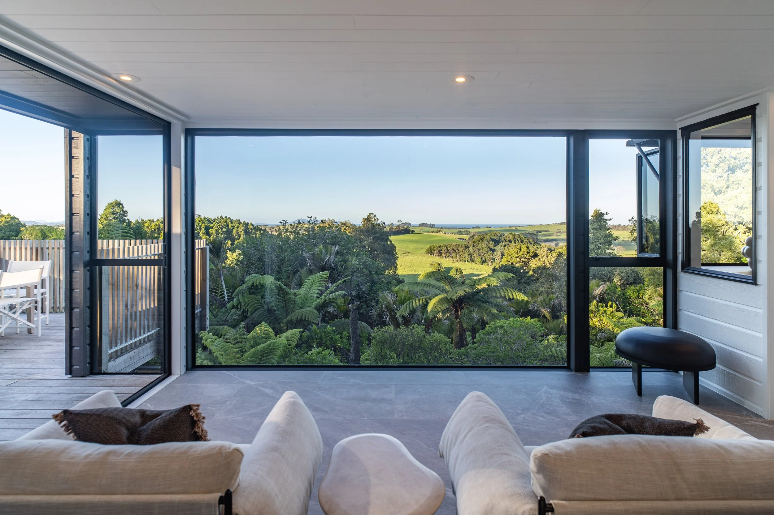 Living room with large glass doors showing green landscape with trees and hills in the distance, white ceiling with recessed lighting, and a beige sofa with pillows.