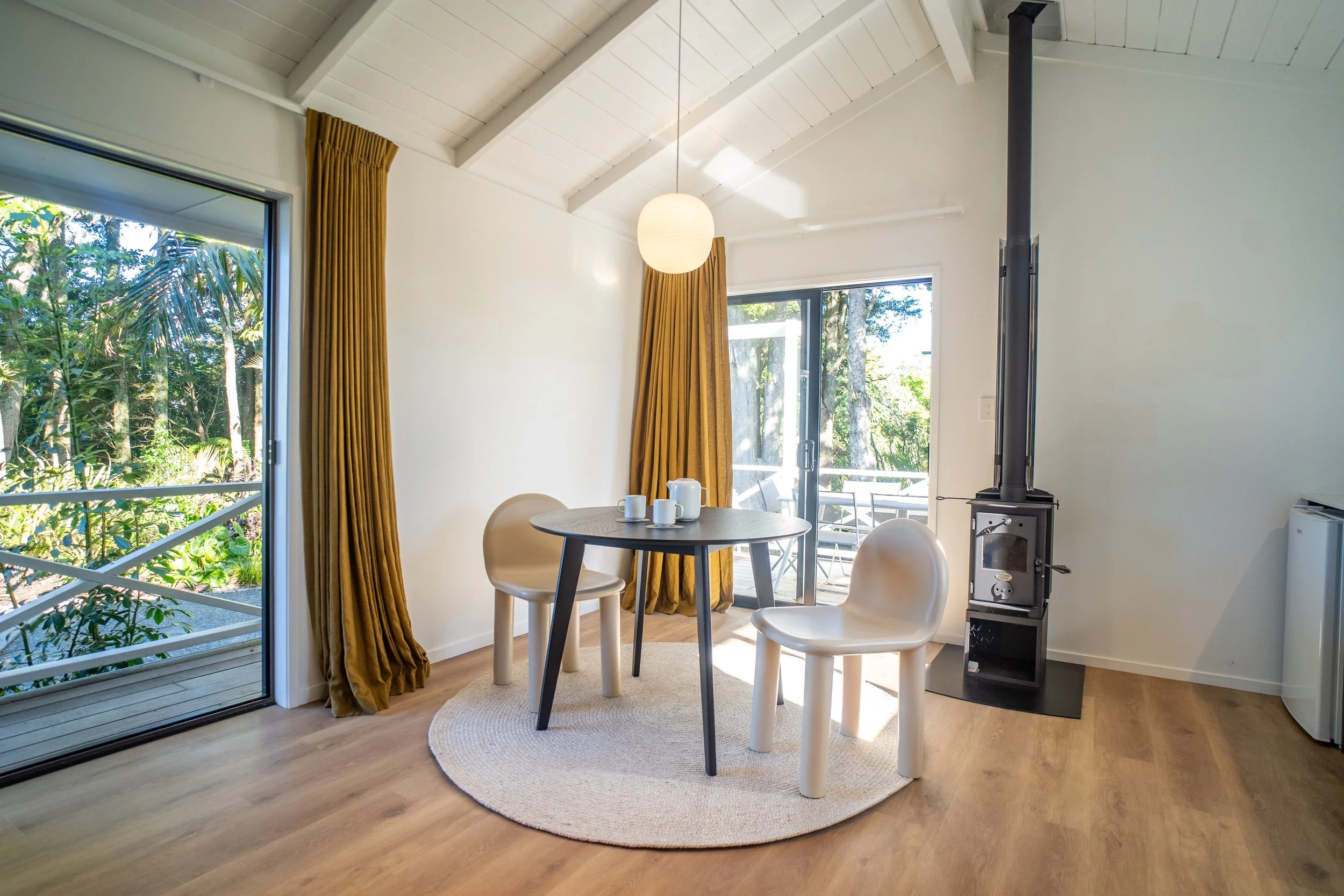 A bright dining area with large sliding glass doors leading to an outdoor deck, gold curtains, a small round table with white cups, two beige chairs, and a wood stove in the corner.