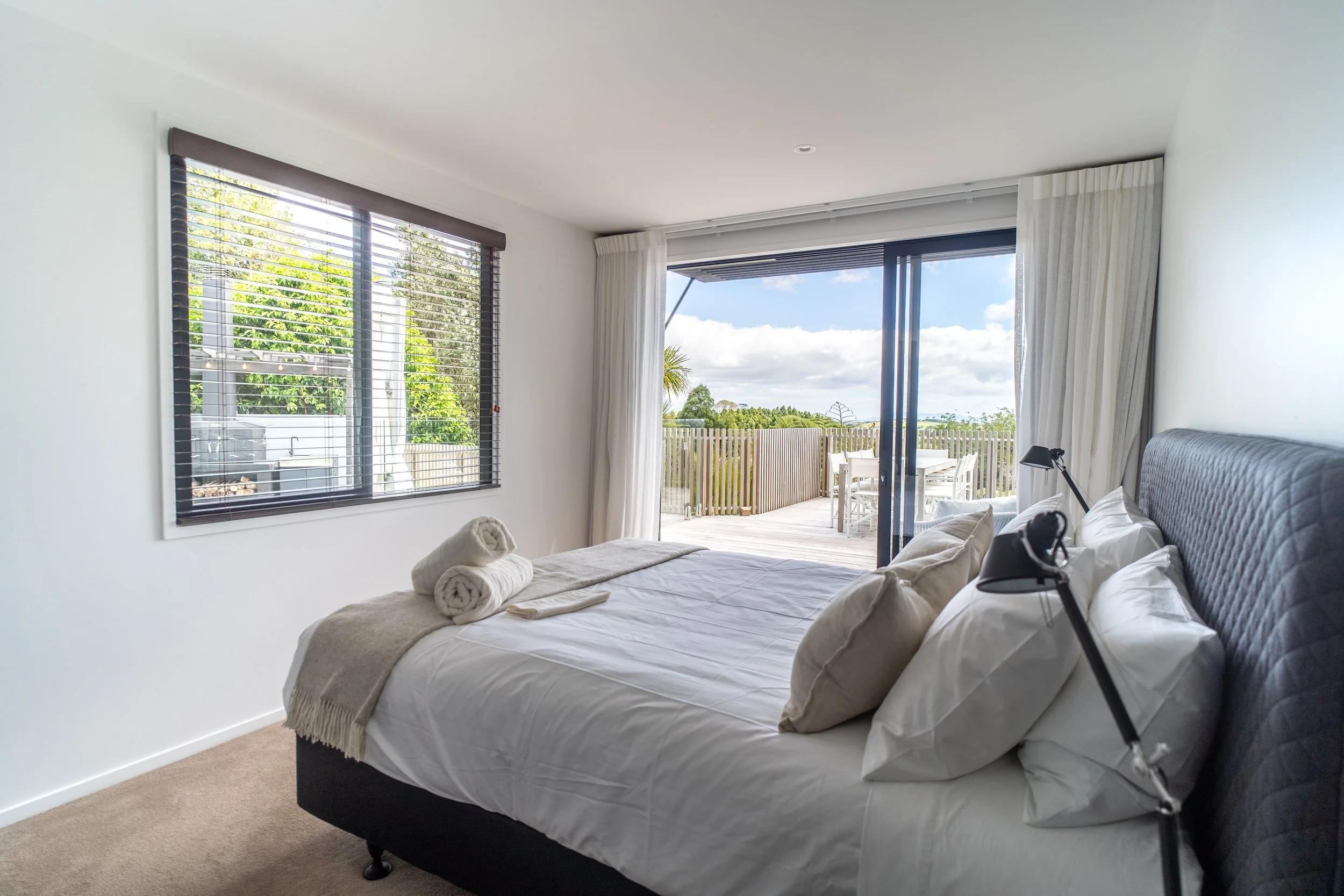A bedroom with a bed covered in white linens and pillows, two rolled towels on top, a window with blinds, and sliding glass doors leading to an outdoor patio with a dining table and chairs, overlooking trees and a blue sky with clouds.