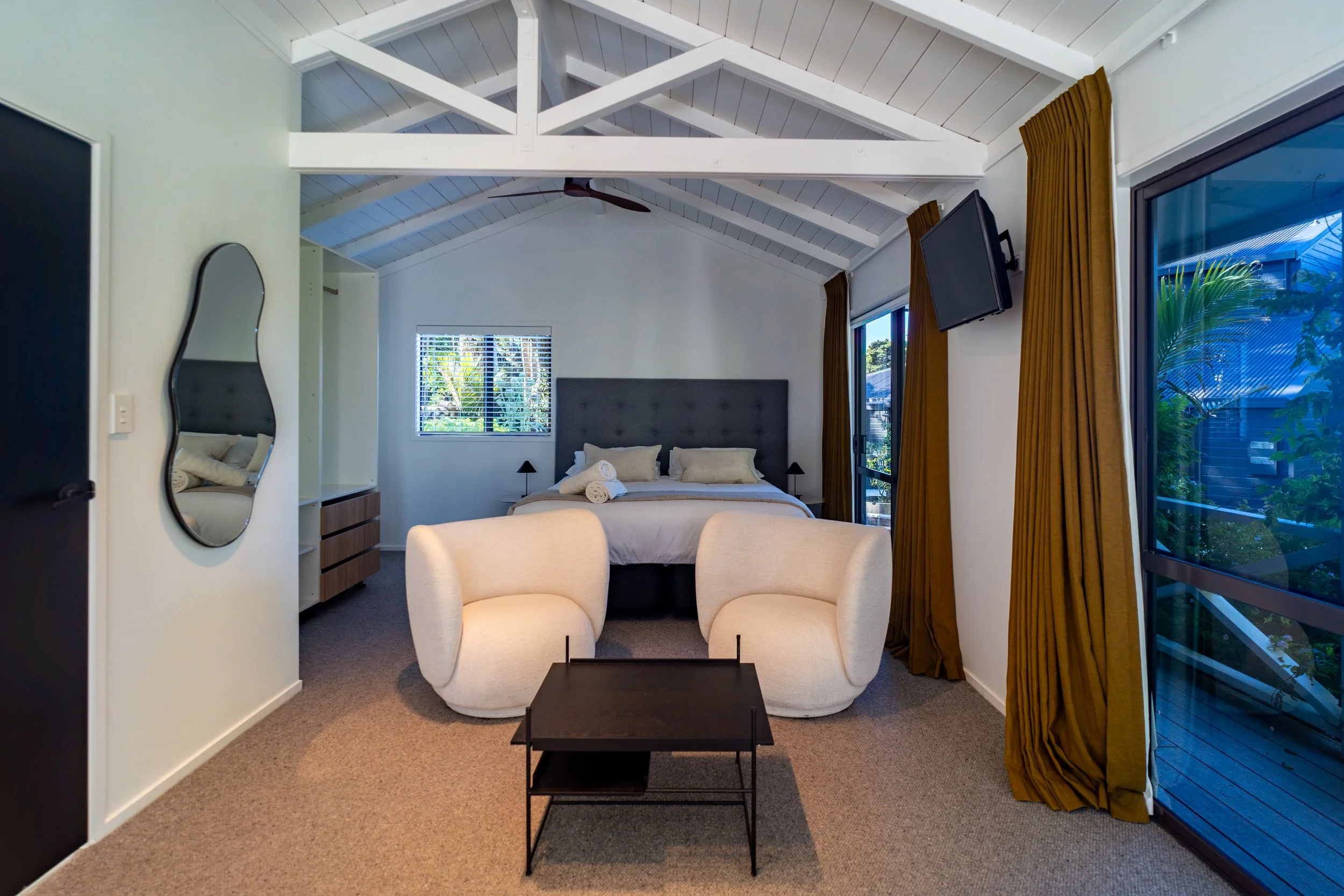 A cozy bedroom with a white vaulted ceiling, a bed with beige bedding, a gray headboard, and two small black nightstands with lamps. Two white armchairs and a black coffee table are in the foreground. Large windows with brown curtains and a sliding g