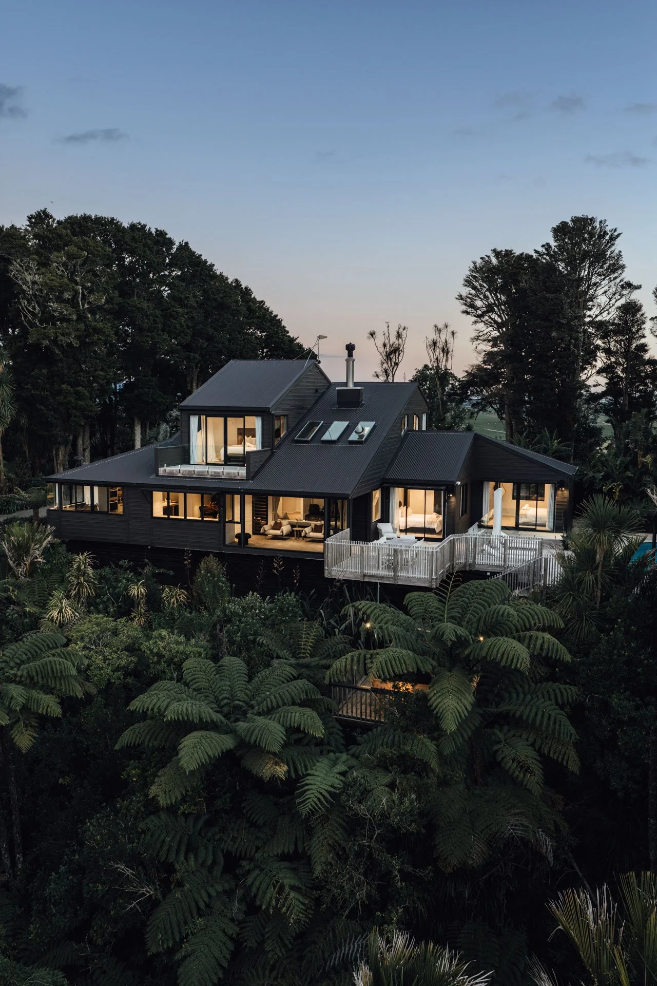 A modern house with large windows and a balcony, surrounded by lush green trees and foliage, at dusk.