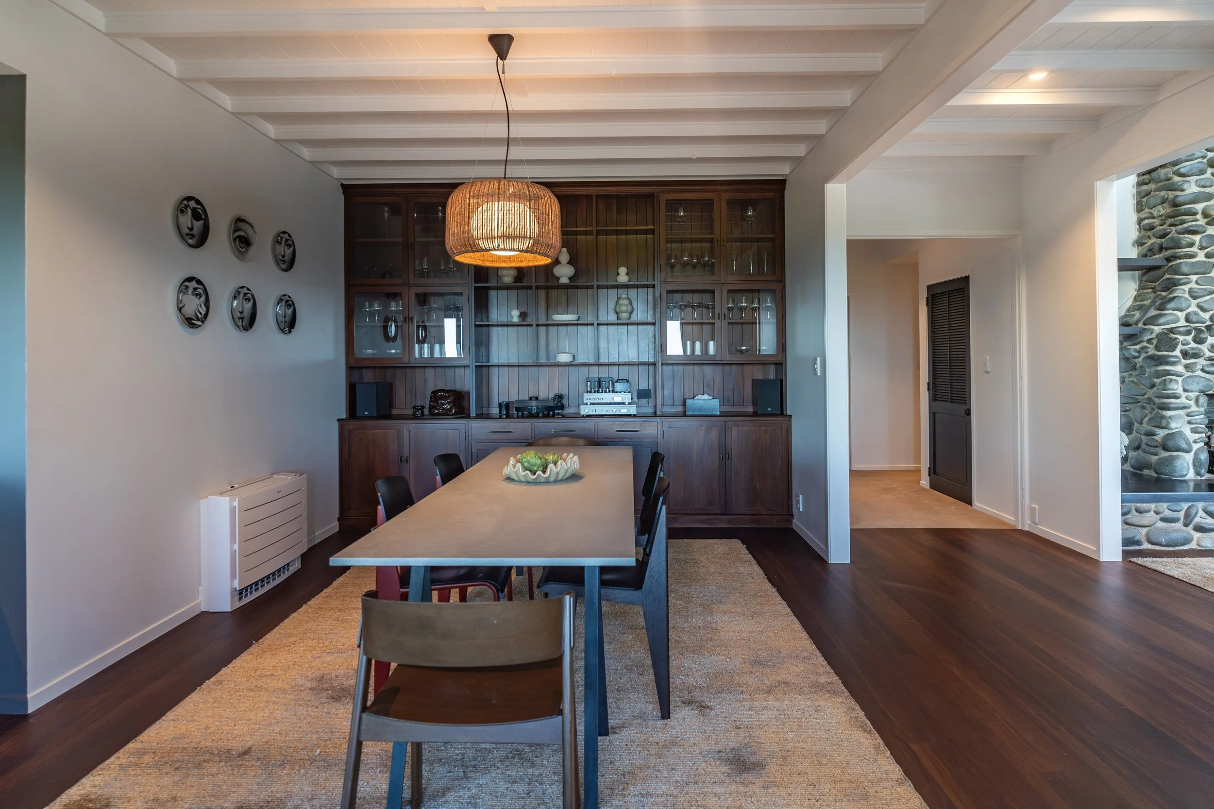A dining room with a wooden table and black chairs, a built-in wood cabinet with glass doors, a hanging wicker light, wall art of black and white faces, a stone accent wall, and a beige rug.