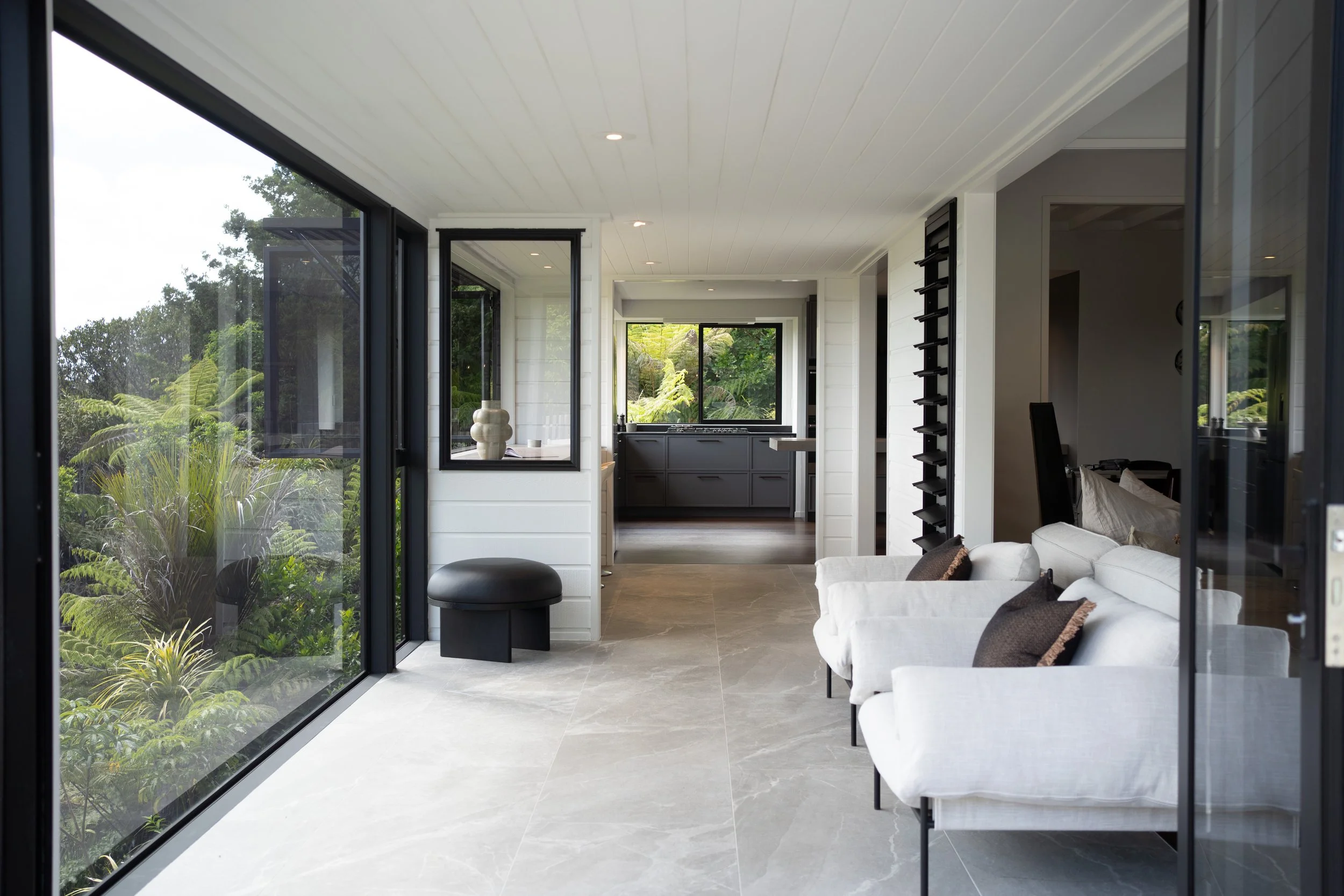 Modern living room with large glass windows overlooking green outdoor garden, white sofa with pillows, TV, and kitchen area in background.