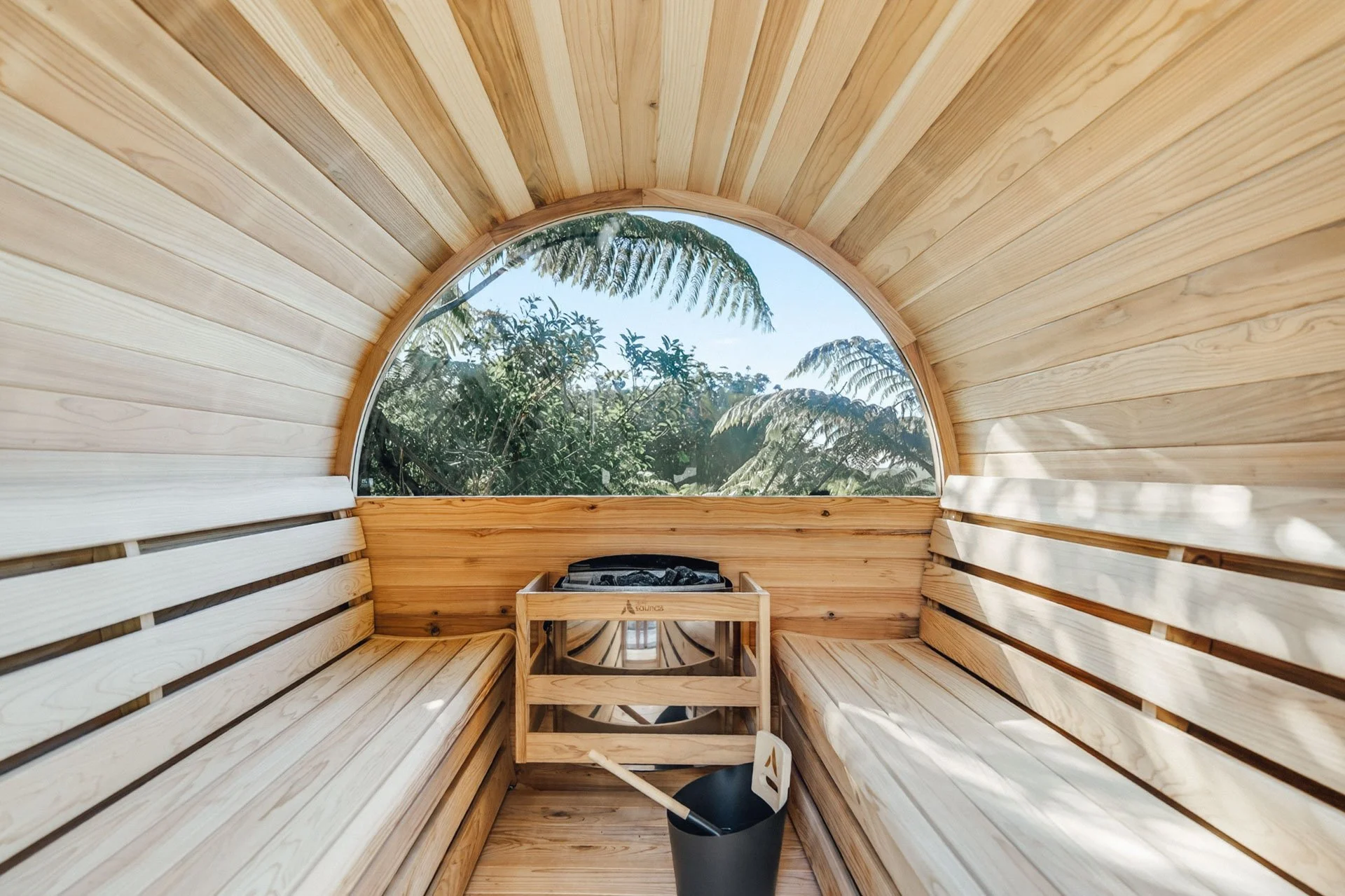 Inside a wooden sauna with slatted walls and benches, a black bucket with a ladle, and a view of trees through a round window.