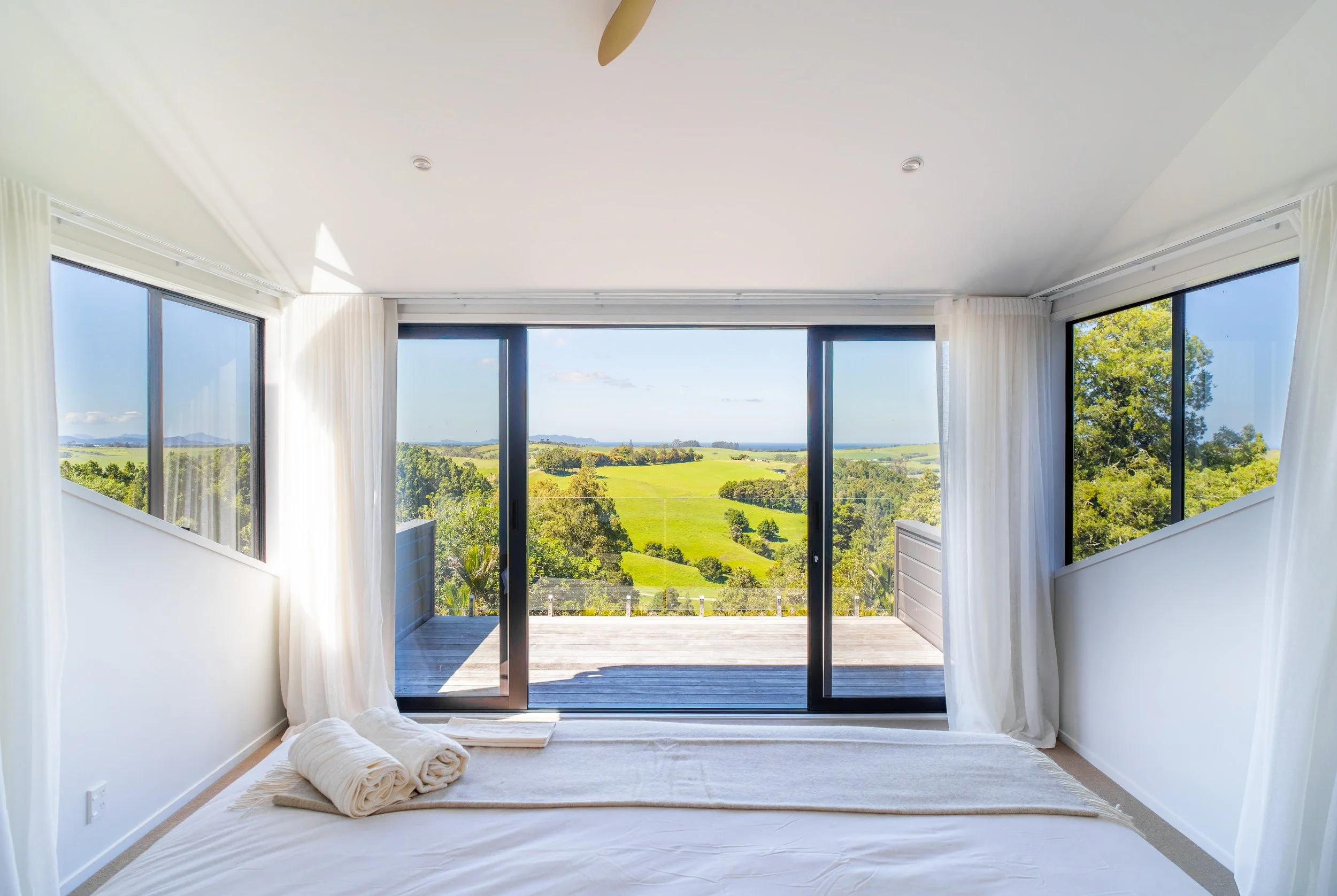 A bedroom with large sliding glass doors leading to a balcony, overlooking a lush green landscape and blue sky, with white curtains on either side.