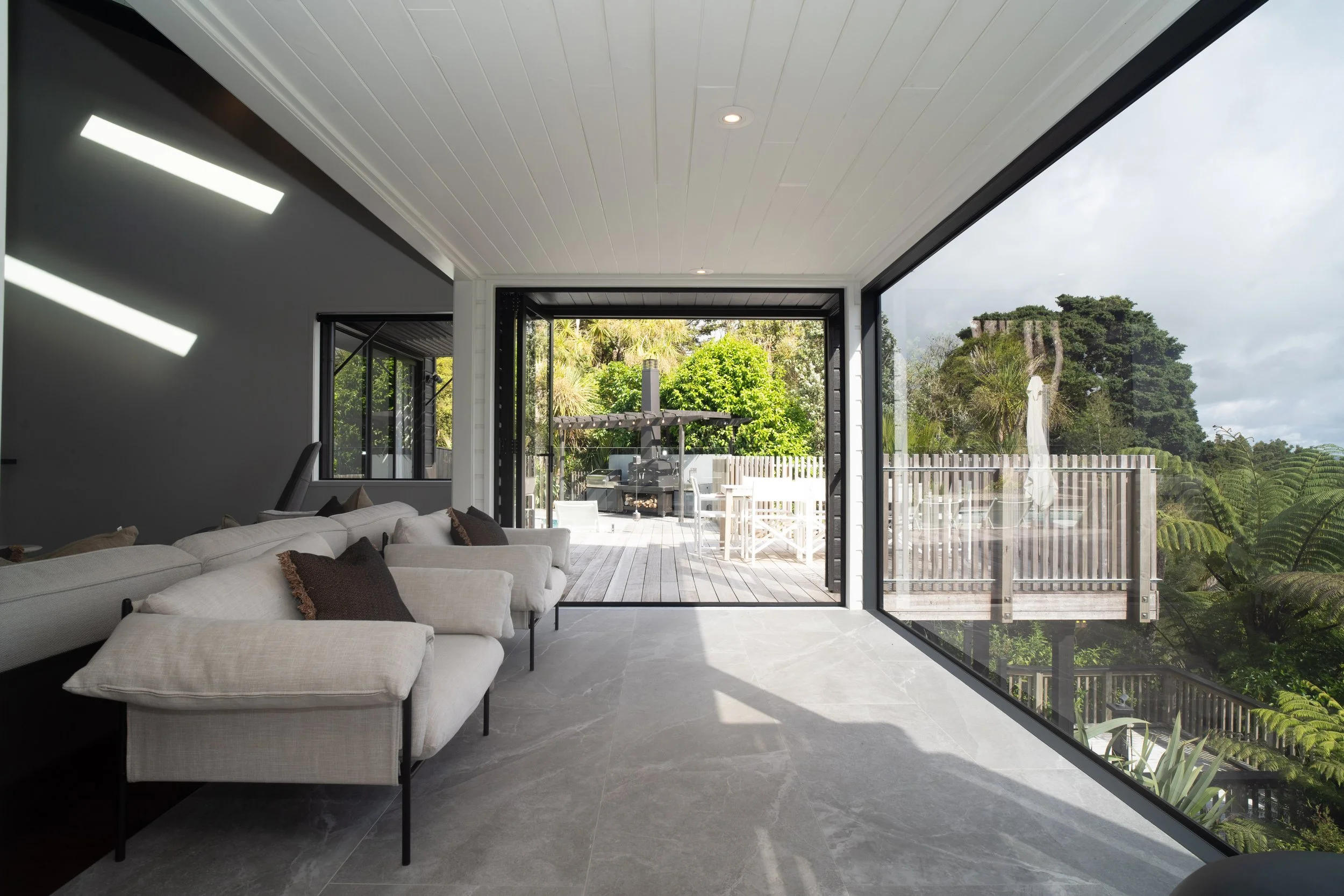 Contemporary living room with white sofa and brown pillows, large glass sliding doors open to a wooden deck outdoor patio with dining table, lush green trees and a hill with a building in the background.
