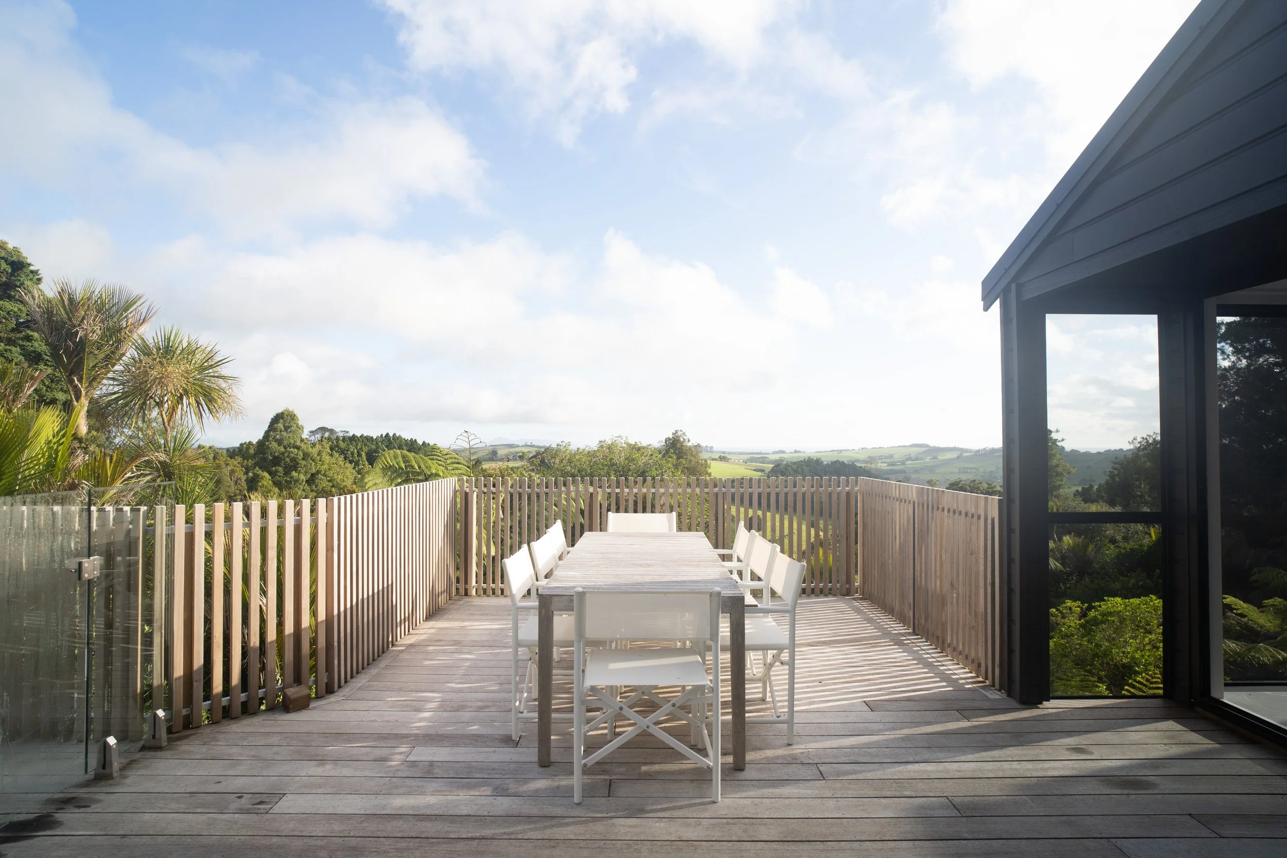 A spacious outdoor balcony with a white dining table and chairs, surrounded by a wooden fence, overlooking green hills and trees under a blue sky with some clouds.