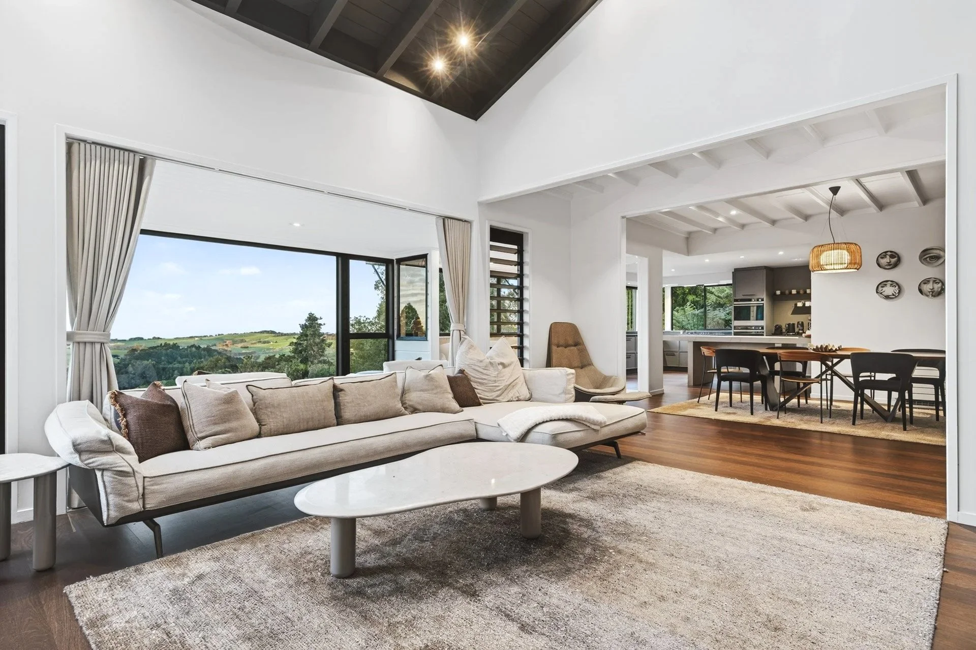 Modern open-plan living room with a large window showing scenic landscape, cream sectional sofa with cushions, white oval coffee table, hardwood floors, area rug, adjacent dining area with black chairs, kitchen with gray cabinets, and wall decorations.