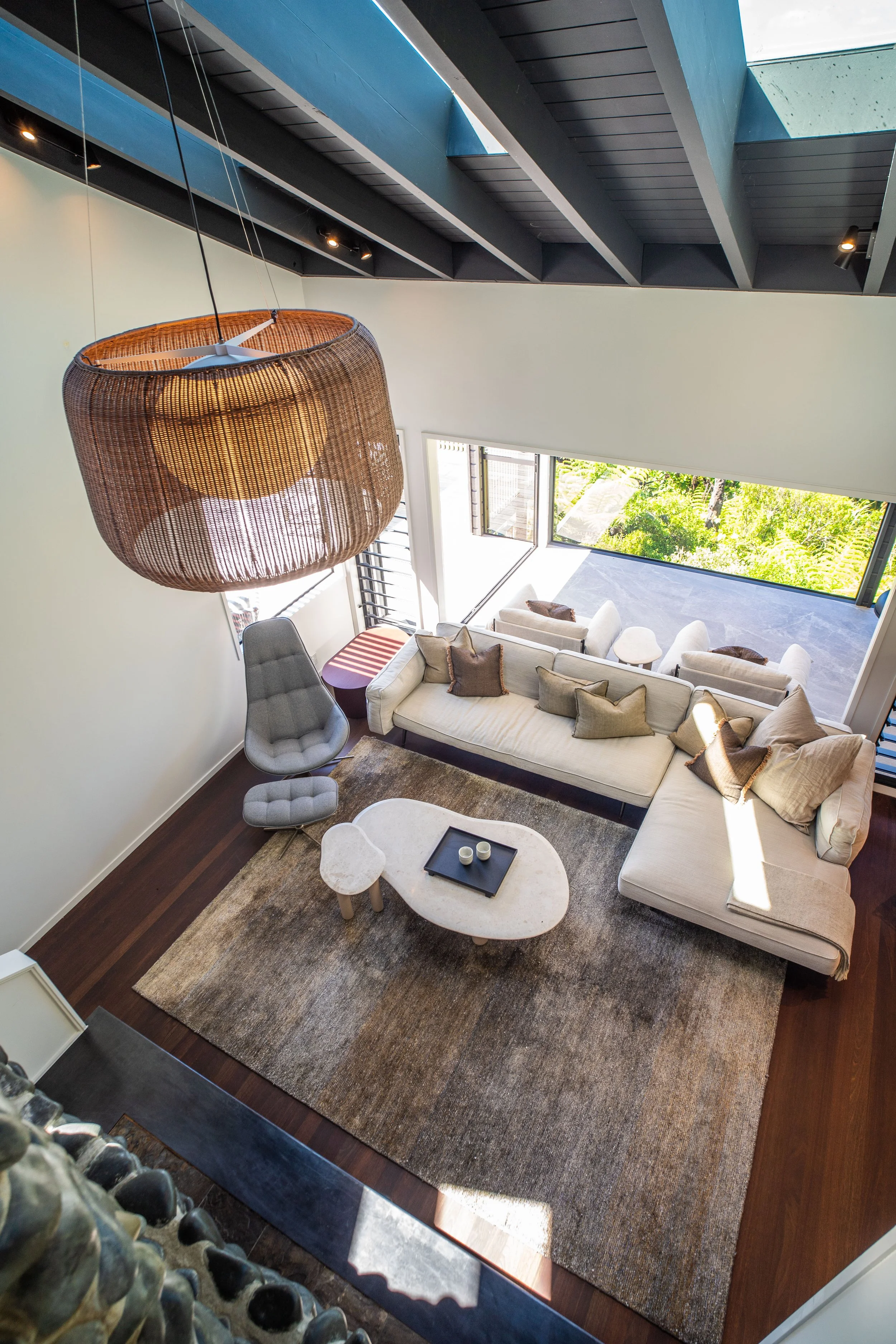 A modern living room with a high ceiling featuring exposed dark beams and a large skylight. The room has a large L-shaped beige sofa with several pillows, a mid-century modern coffee table with a black tray and two cups, a patterned area rug, and a u