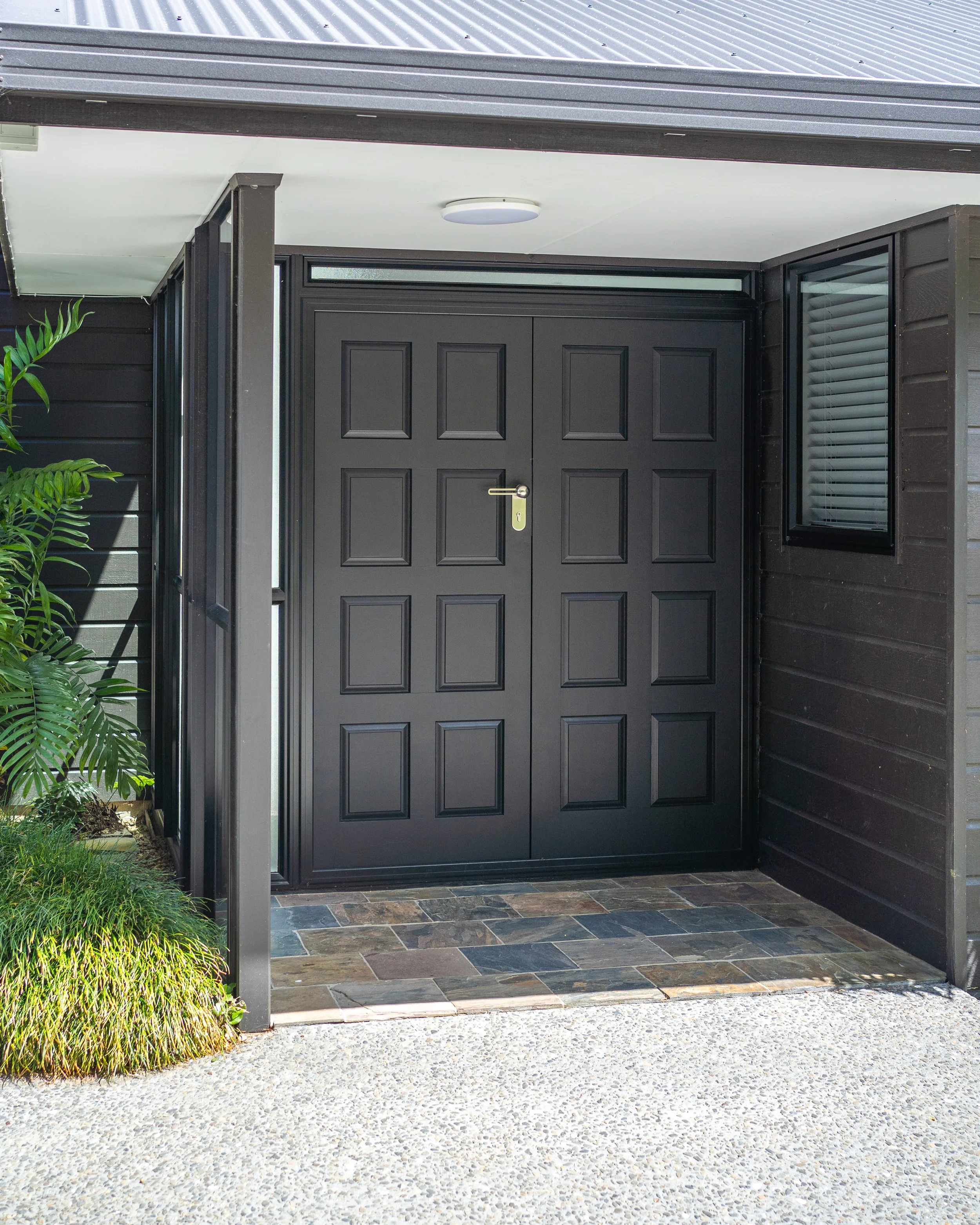 Black front door with rectangular panels, surrounded by black siding, with a window above and to the right, and a round ceiling light fixture above. There are green plants on the left and a tiled floor in front.