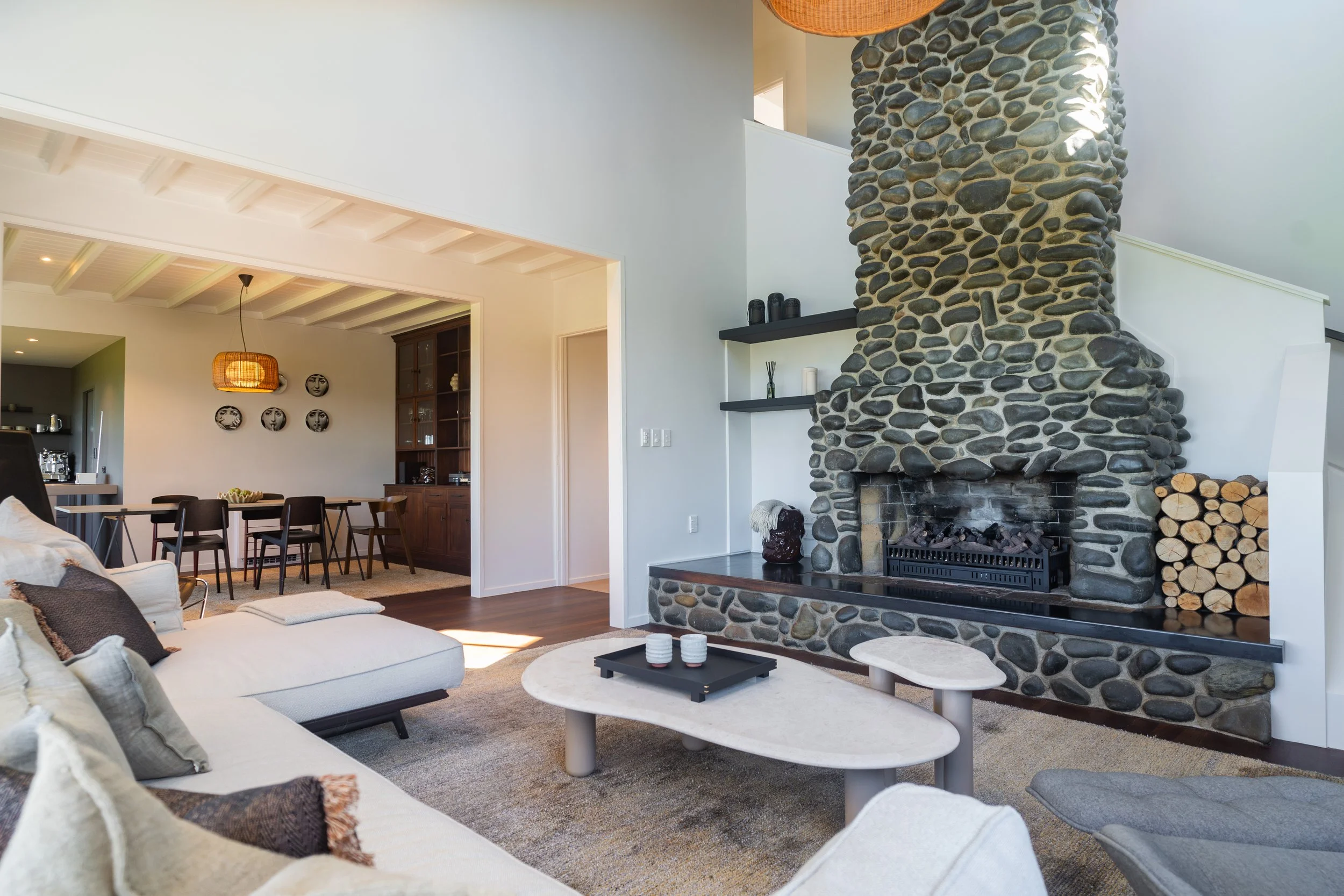Living room with a large stone fireplace, beige sectional sofa with throw pillows, two round coffee tables, and a dining area with a hanging light fixture and wall art.