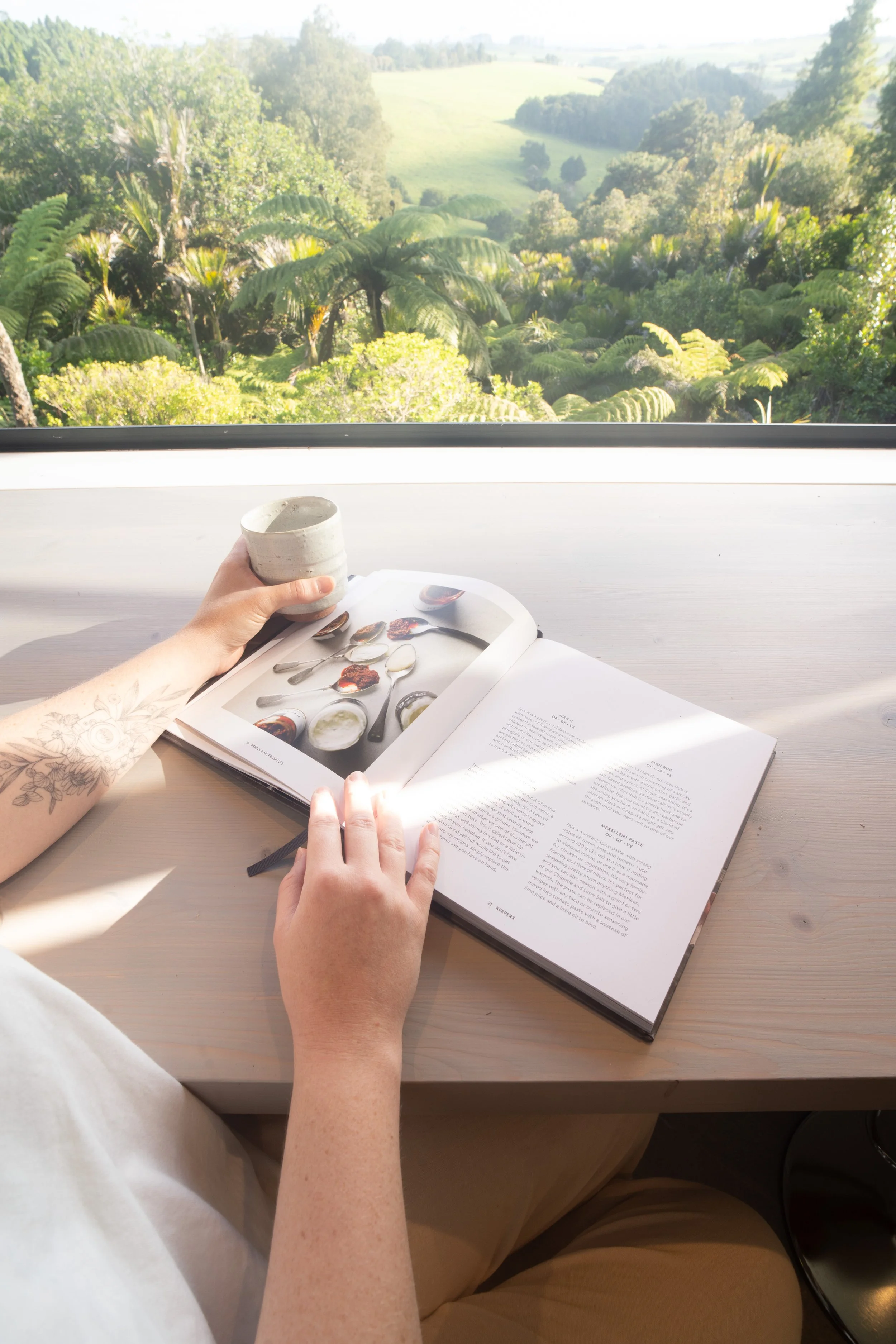 A person sitting at a light-colored wooden table reading a magazine with images of spoons and bowls, holding a small ceramic cup with a light-colored drink, with a view of lush green trees and rolling hills outside a window in the background.