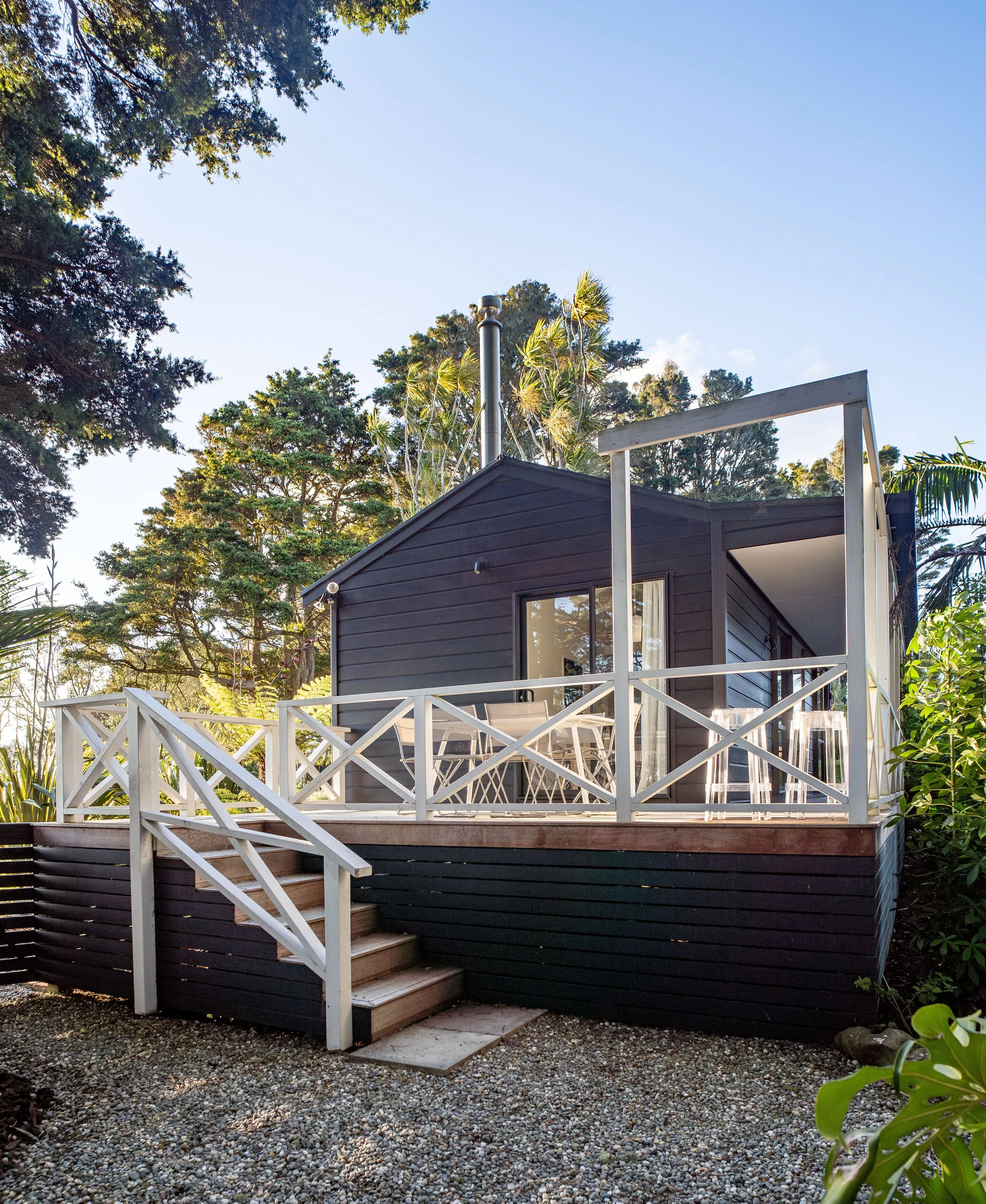 A modern black house with a white wooden deck and stairs, trees, and a clear blue sky in the background.