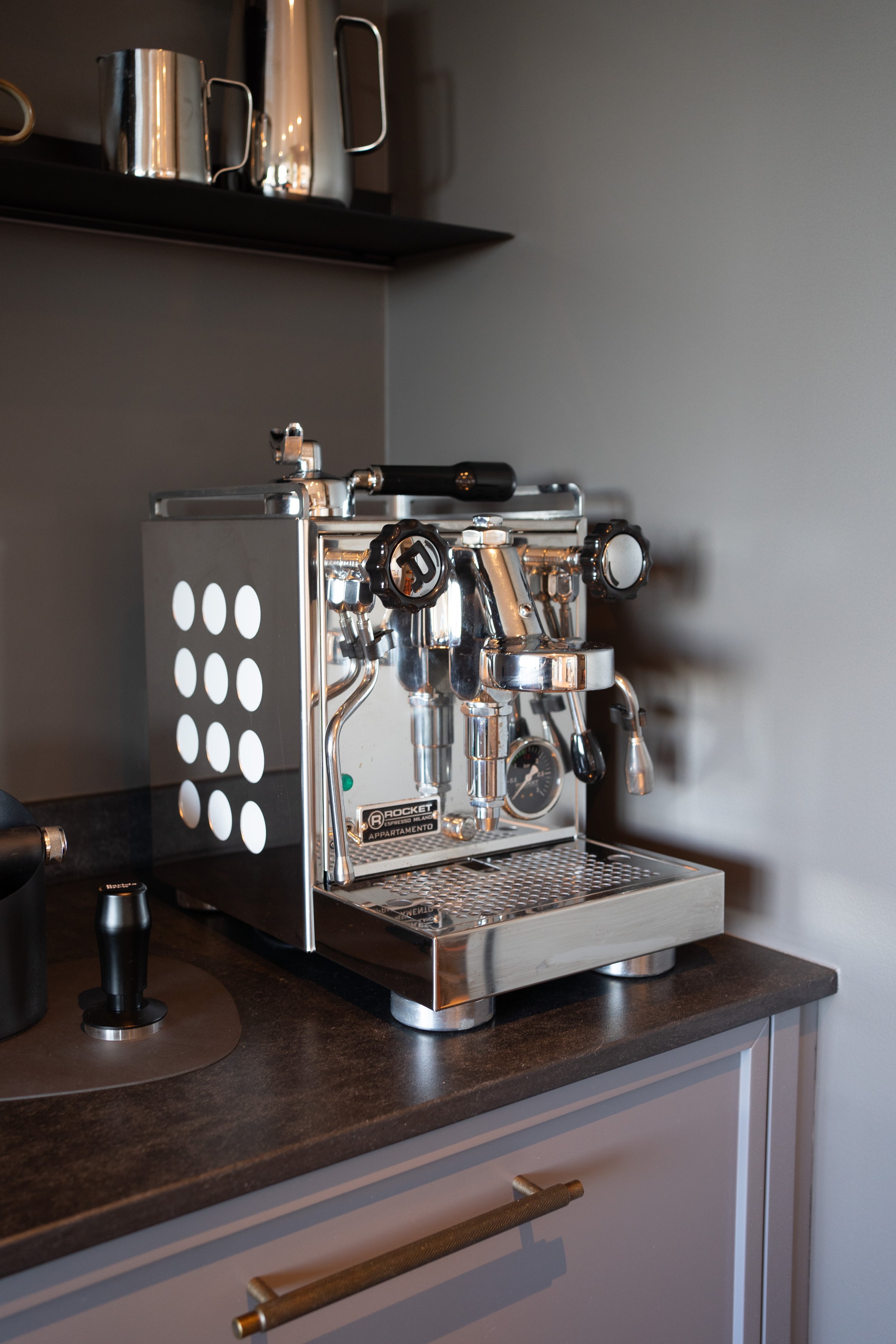A shiny, silver espresso machine on a dark countertop with a drawer below. The machine has multiple knobs, a gauge, and a steam wand. There is a black object and a black round object on the counter next to it. A gray wall is in the background and a f