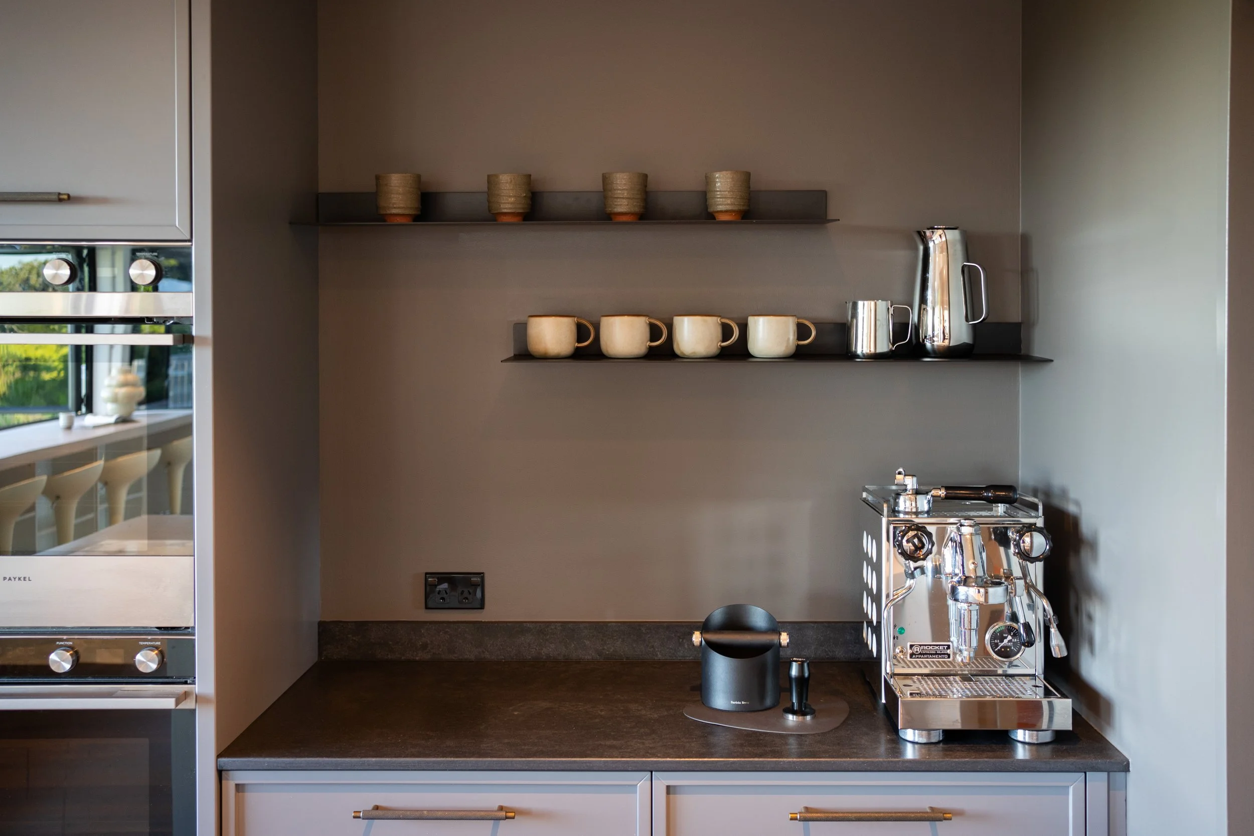 A kitchen countertop with a silver espresso machine on the right, a black grinder in the center, a power outlet on the wall, and two shelves with cups and mugs. The left side shows part of a refrigerator and a window with a view outside.