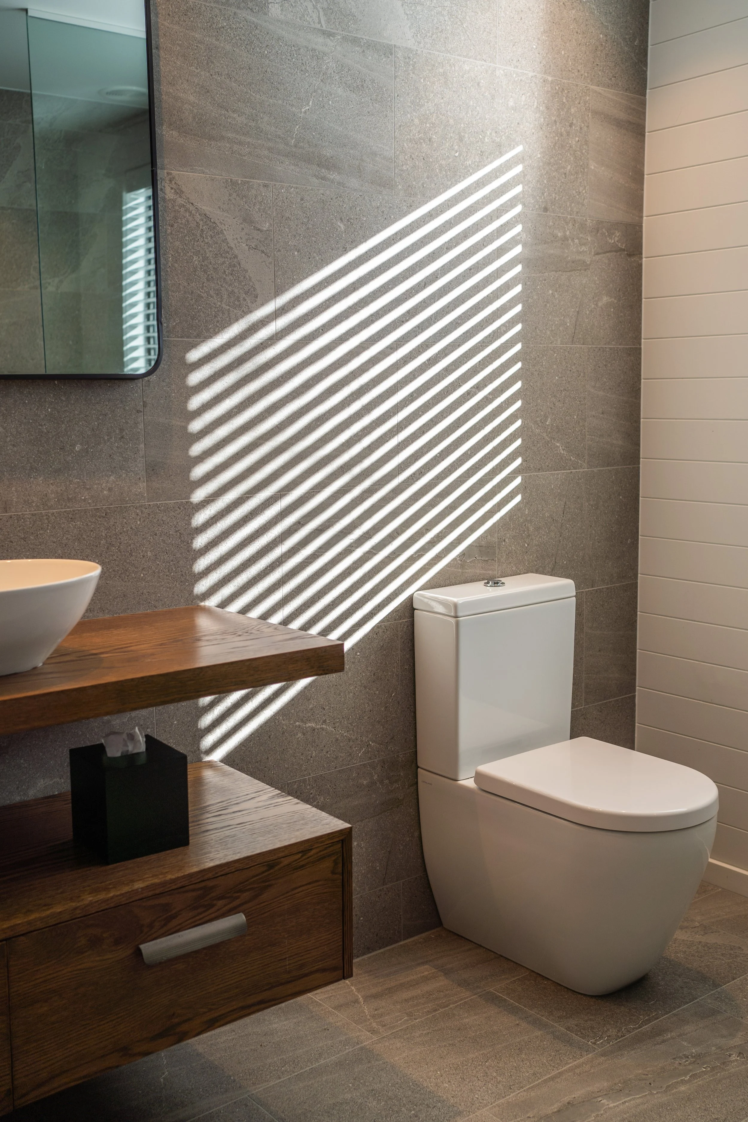 A modern bathroom with gray tiled walls and floor, a white toilet, a wooden vanity with a white bowl sink, a black tissue box, and a mirror reflecting a window with blinds, with sunlight creating diagonal shadows on the wall.