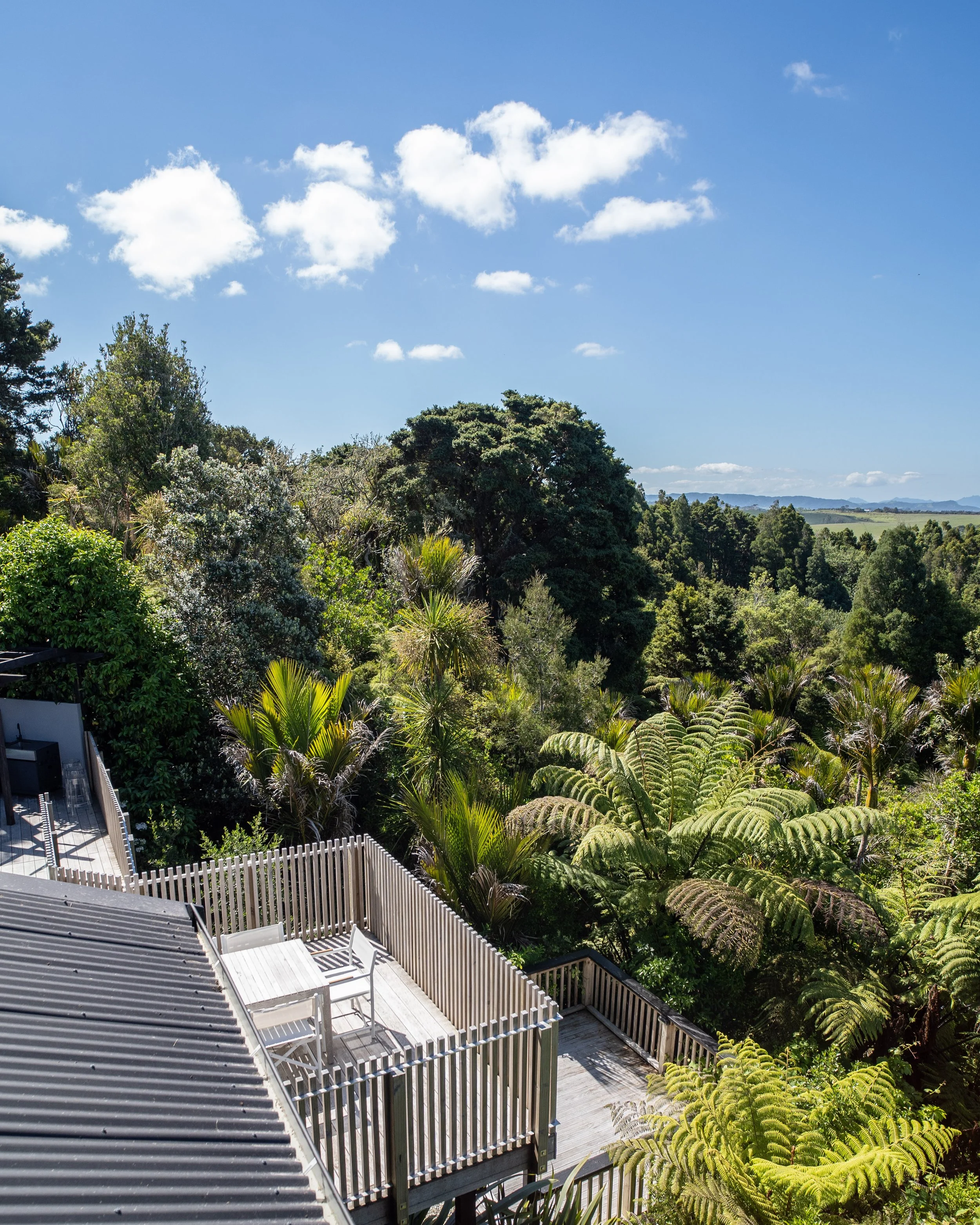 A view of a backyard deck with white chairs and a railing, overlooking lush green trees and a distant landscape with hills under a partly cloudy blue sky.