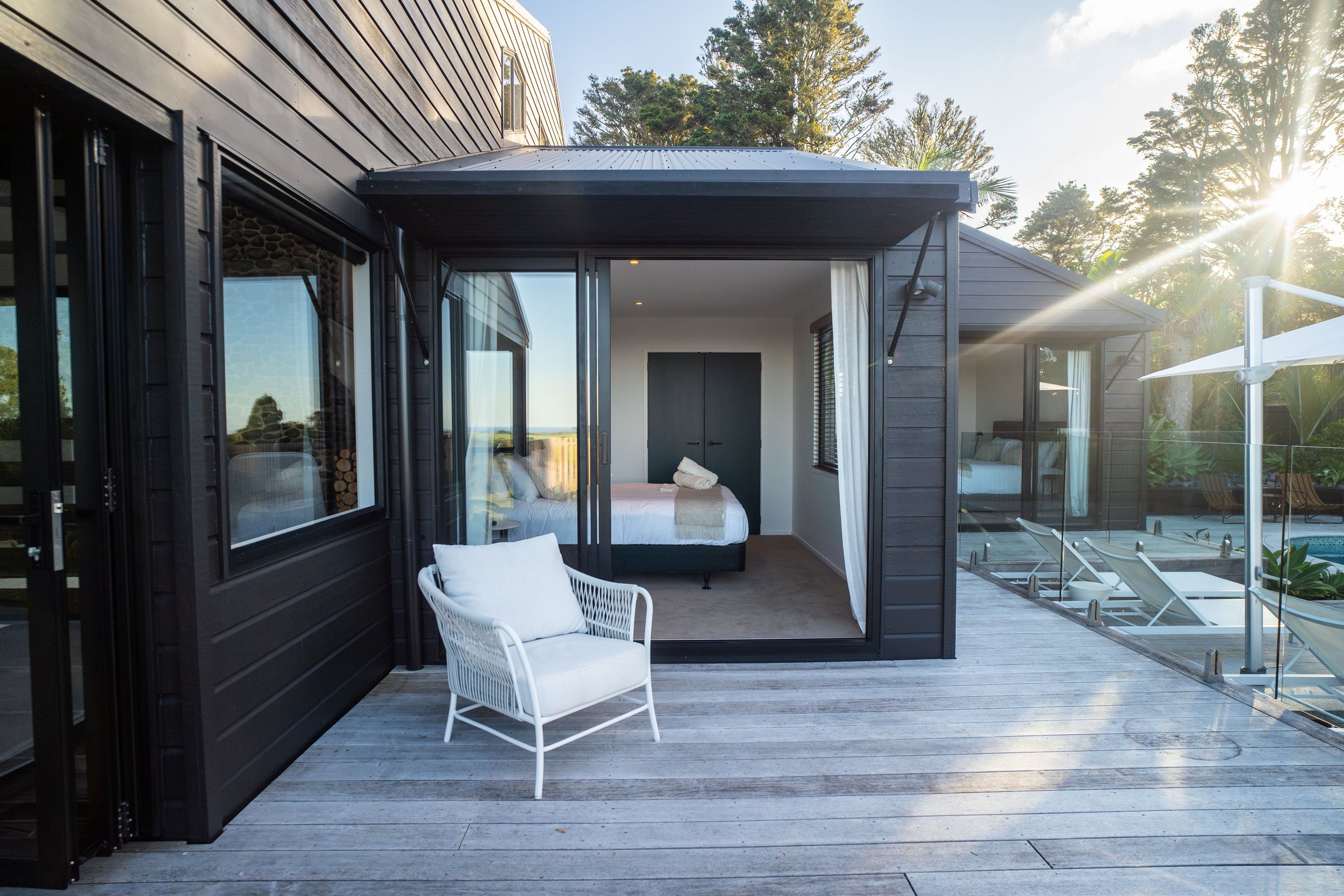 A modern house with a deck, glass sliding doors, and an outdoor chair, surrounded by trees and sunlight.