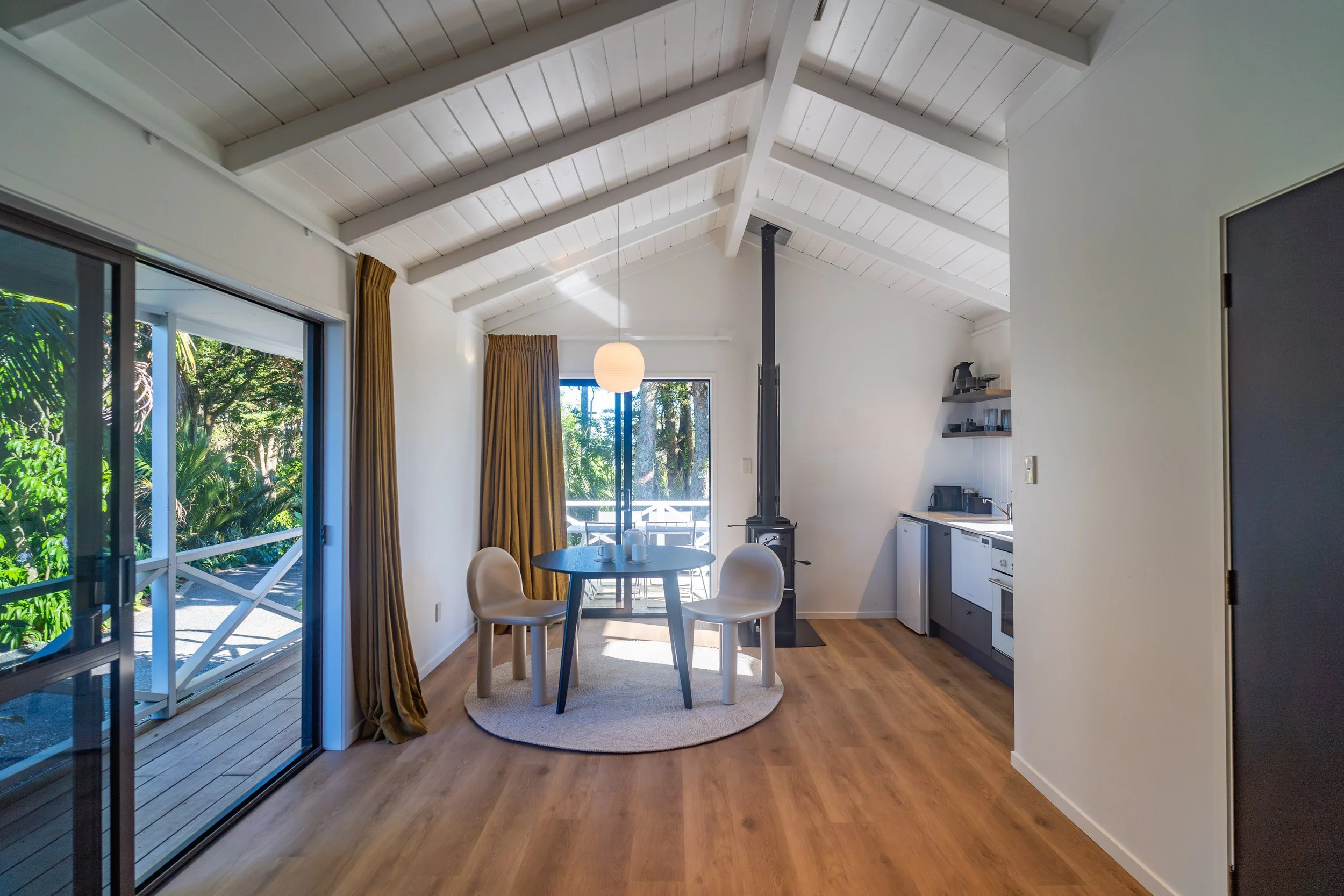 Interior of a modern home with a high white wood-beamed ceiling, sliding glass doors leading to a deck with trees outside, a round dining table with four beige chairs, a paper lantern hanging from the ceiling, and a small kitchen area with a countert