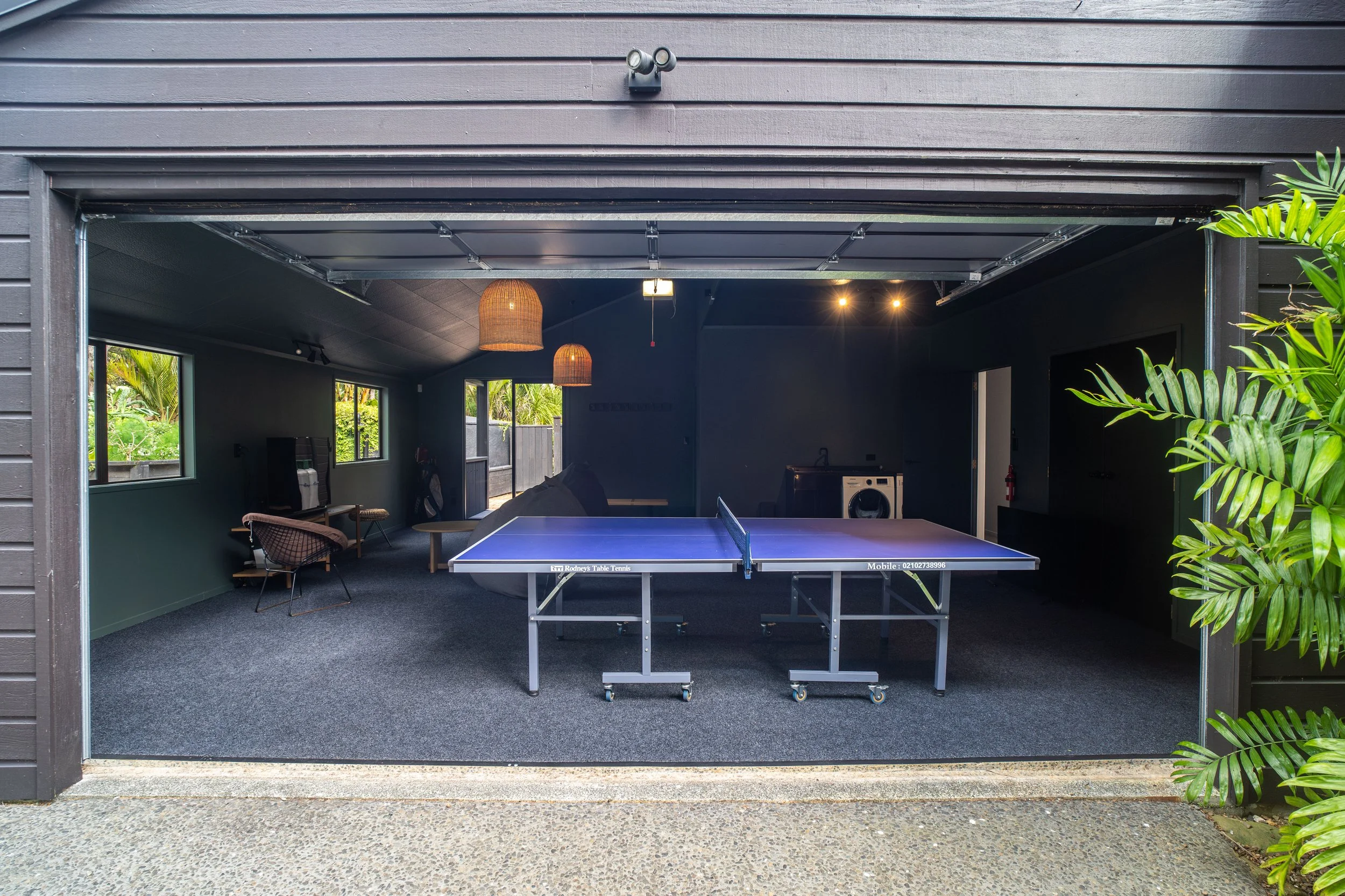Open garage door revealing a game room with a ping pong table, a wicker hanging lamp, a chair, windows showing greenery outside, and indoor plants on the right.