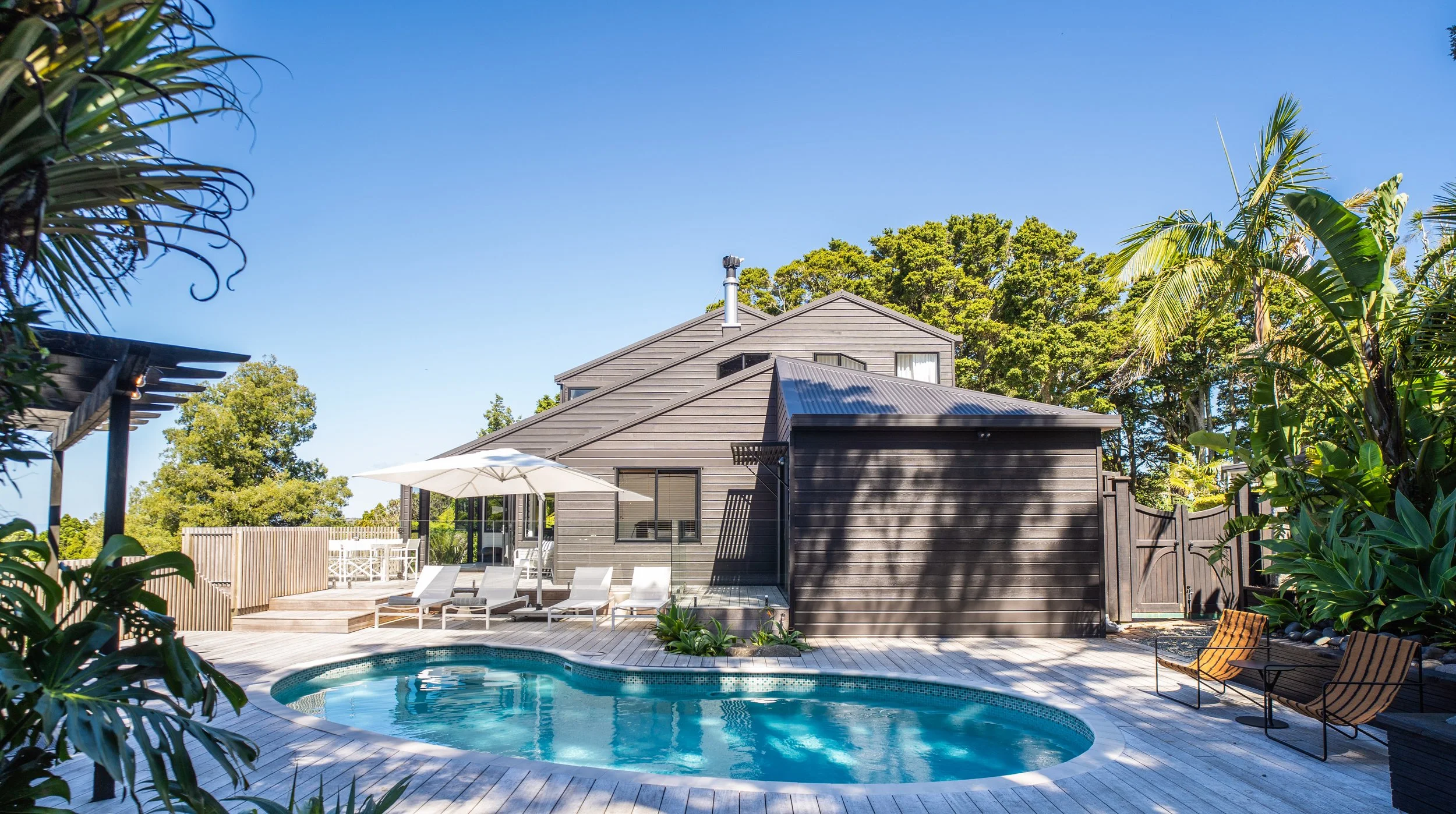Backyard with swimming pool, modern house, outdoor chairs, umbrella, surrounded by lush greenery and trees, under a clear blue sky.