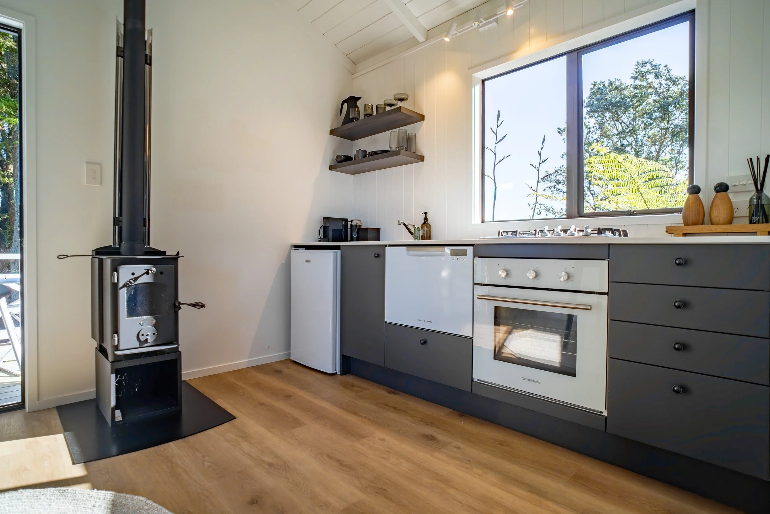 Modern kitchen with large window, gray cabinetry, white walls, and wooden floor, with a wood stove in the corner.