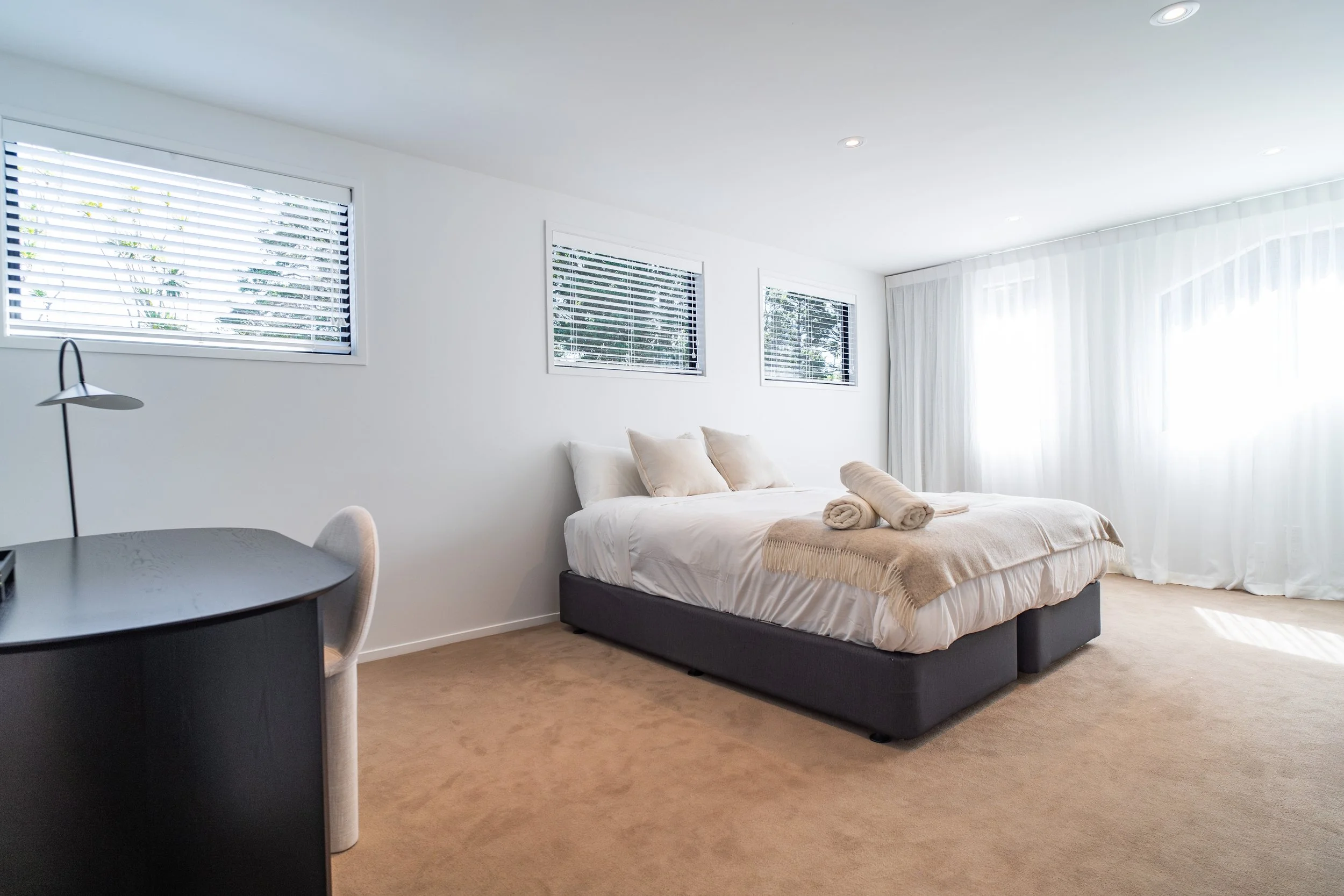 Bright bedroom with large windows, a bed with white linens and rolled towels, a modern desk with a chair, and a white curtain on the right side.