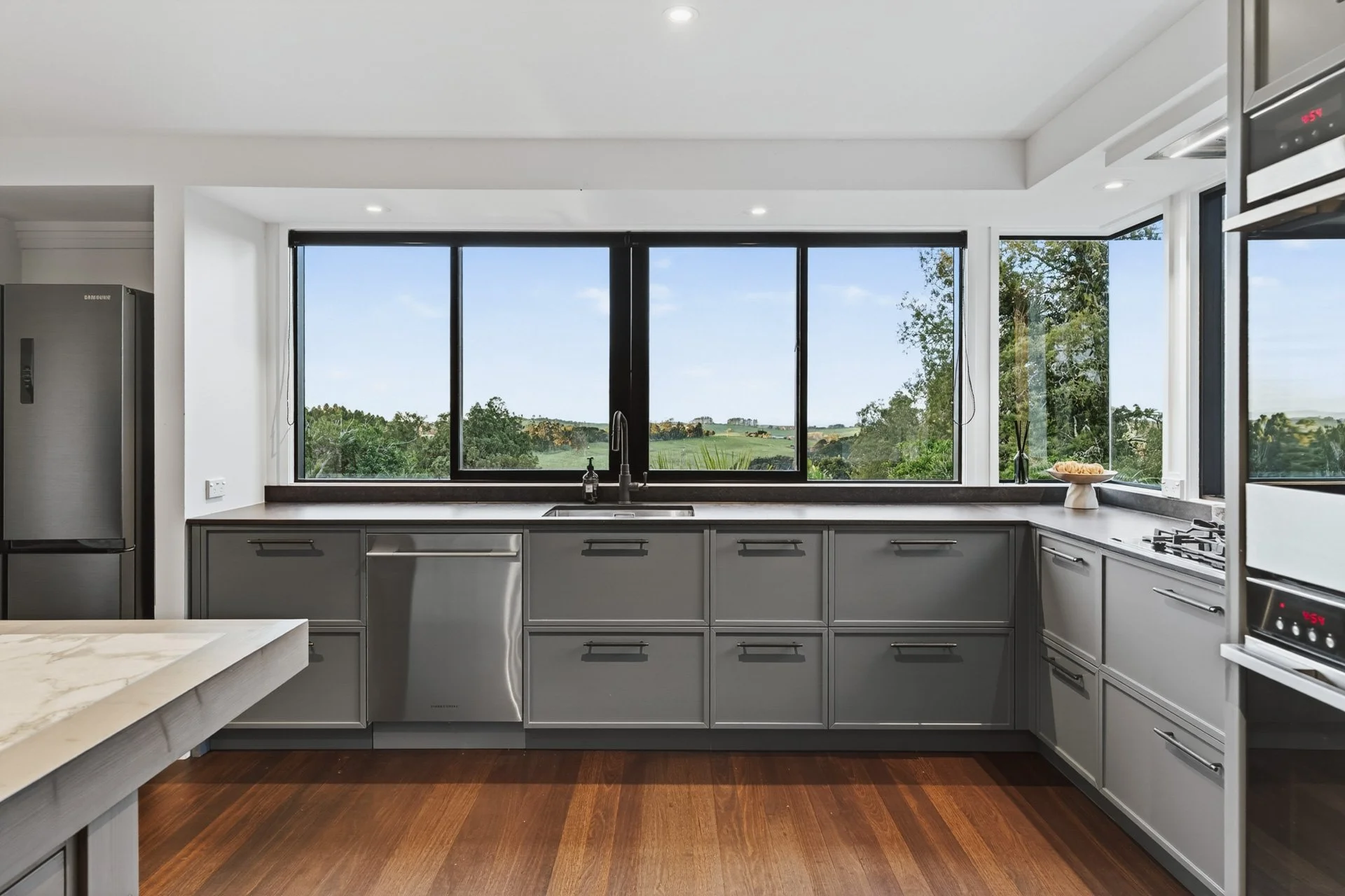 Modern kitchen with gray cabinets, large window showing green landscape, stainless steel appliances, dark wood floor, and white walls.