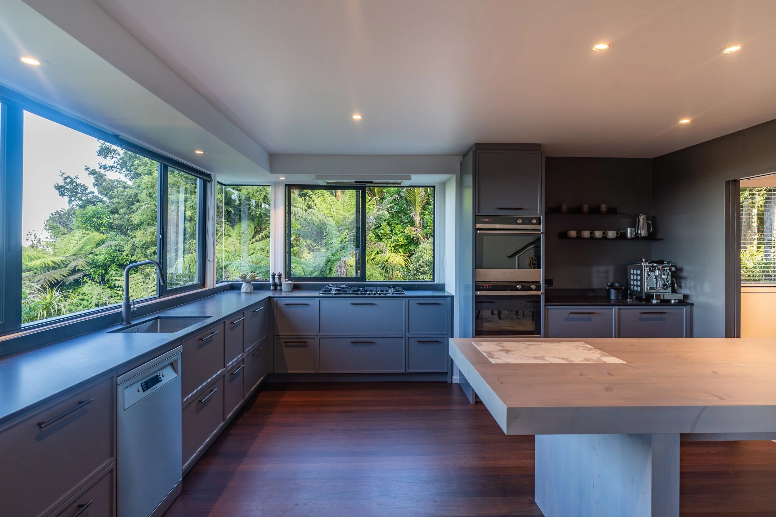 Modern kitchen with large windows showing lush greenery outside, gray cabinets, a black countertop, and built-in appliances.