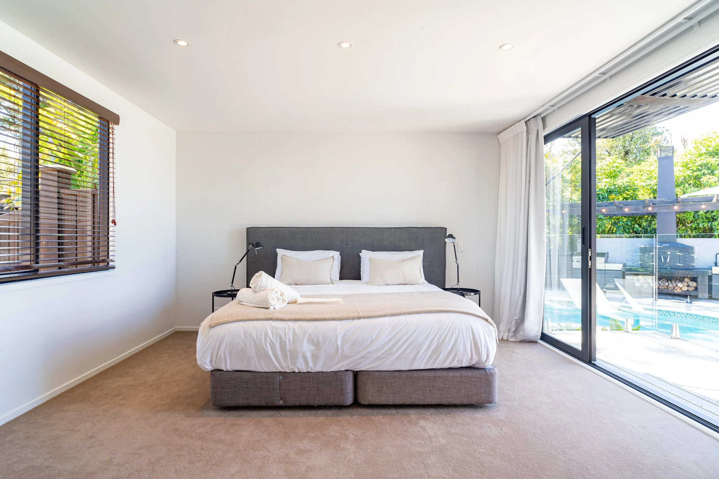 Modern bedroom with large sliding glass doors leading to an outdoor patio and pool, a bed with beige and white bedding, and black side tables with reading lamps.